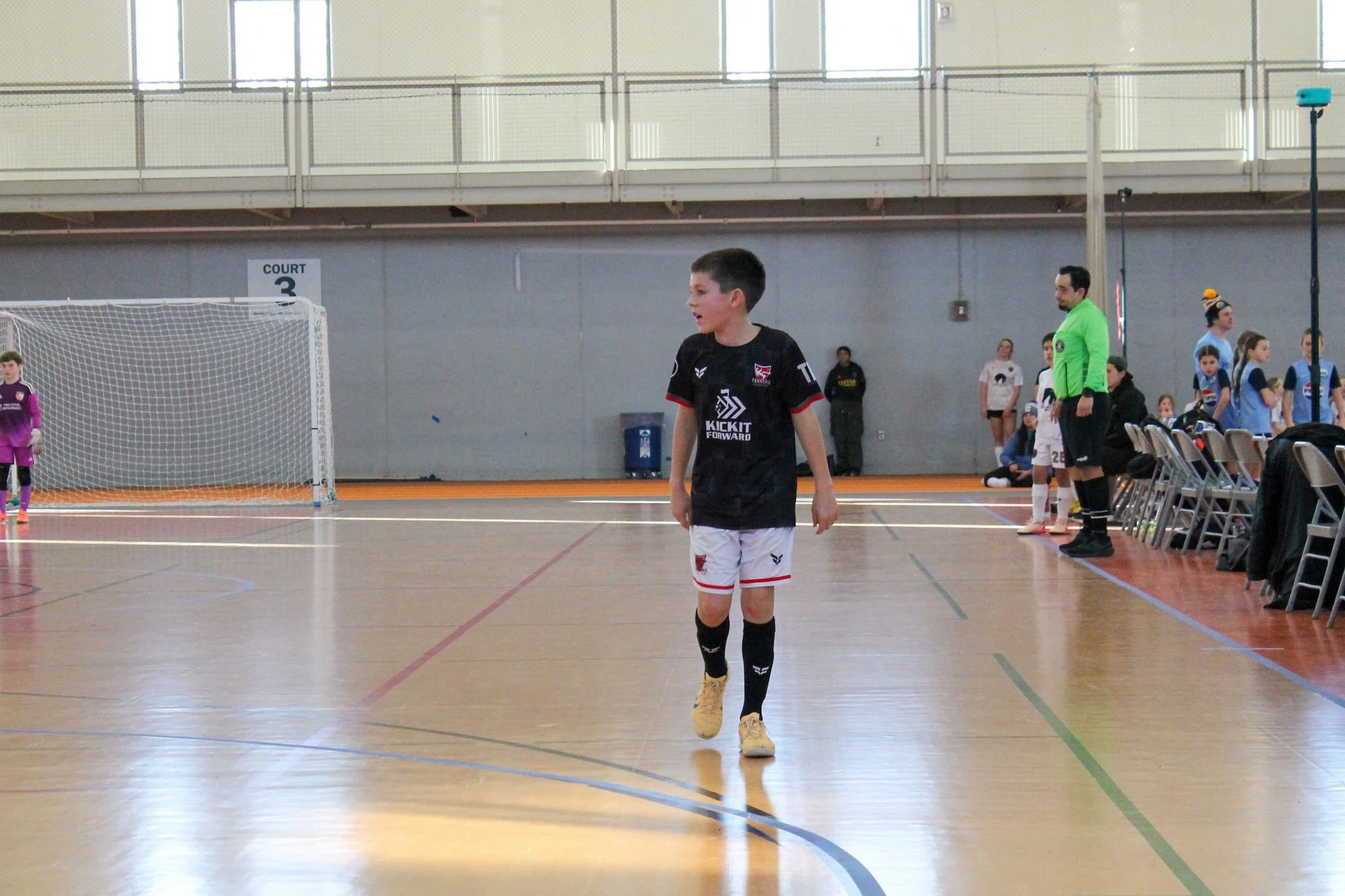 Indoor sports court with young male soccer player in black and white uniform walking across the court, surrounded by other players, coaches, and set up chairs, with goal net and court sign in the background.