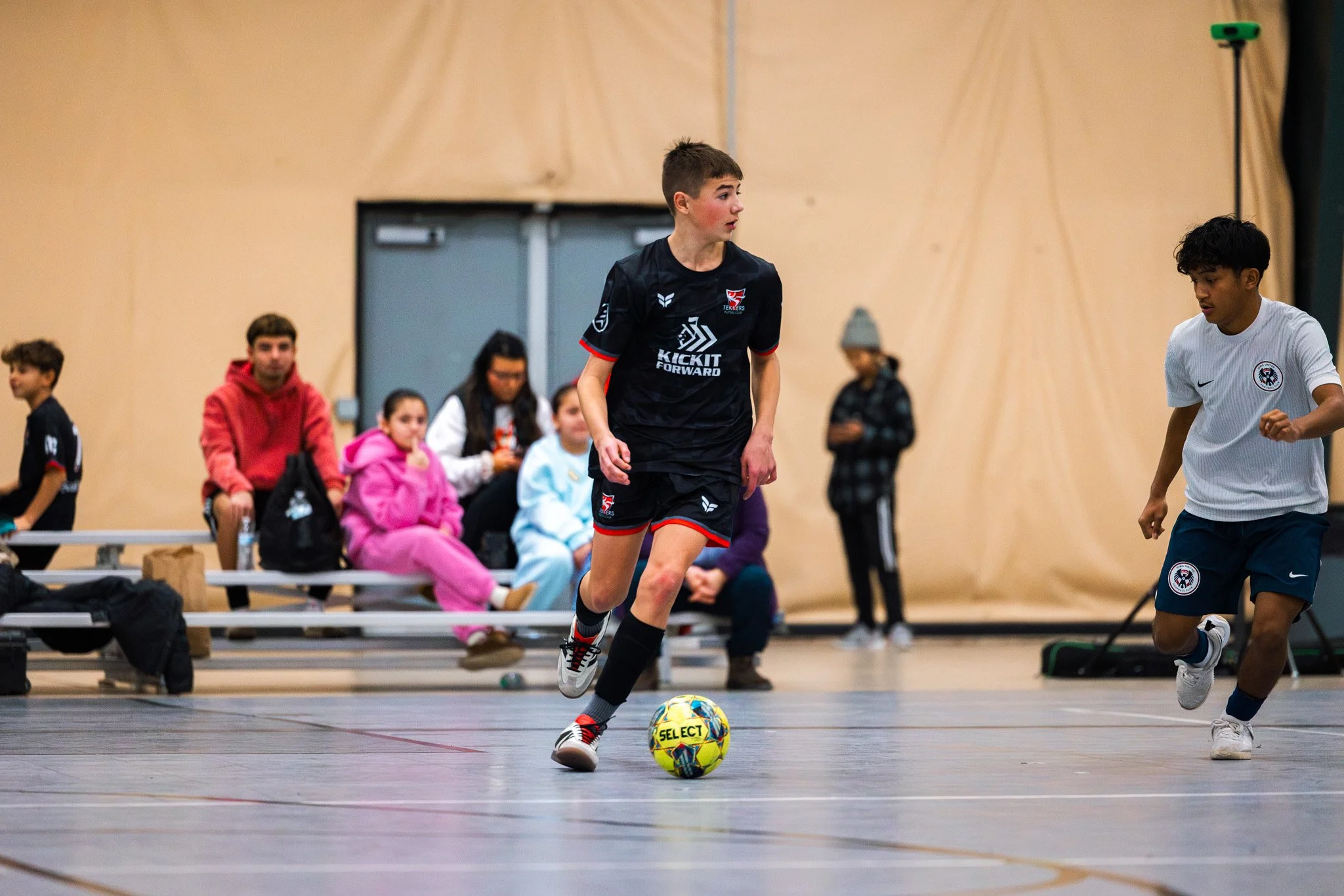 Young boy in black sports uniform dribbling a yellow and blue soccer ball indoors, with spectators sitting on benches in the background.