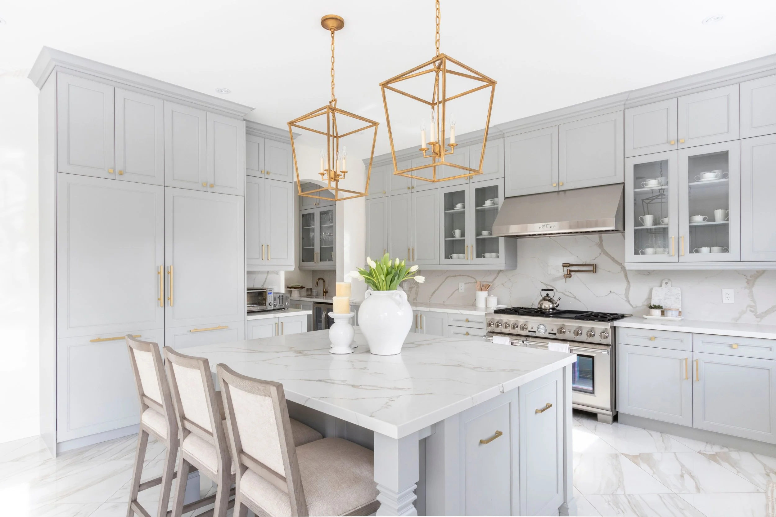 Modern white kitchen with marble backsplash, island with sink, built-in appliances, pendant lights, and a large window.