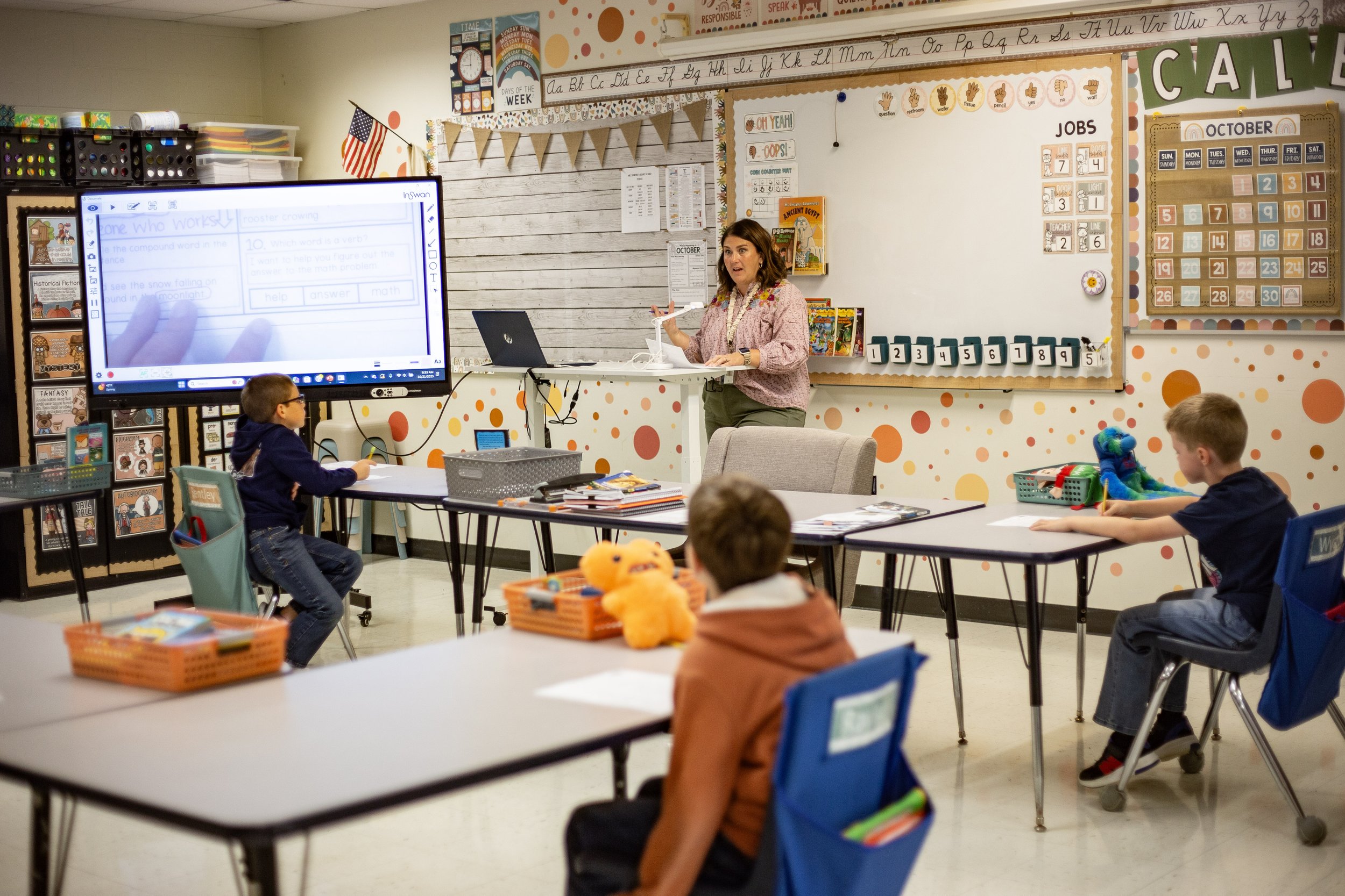 A classroom with students sitting at desks and a teacher standing near a whiteboard. The classroom has maps of the world and flags displayed on the walls. Students are smiling and looking towards the camera, and the teacher is holding a cup. An American flag is visible in the room.