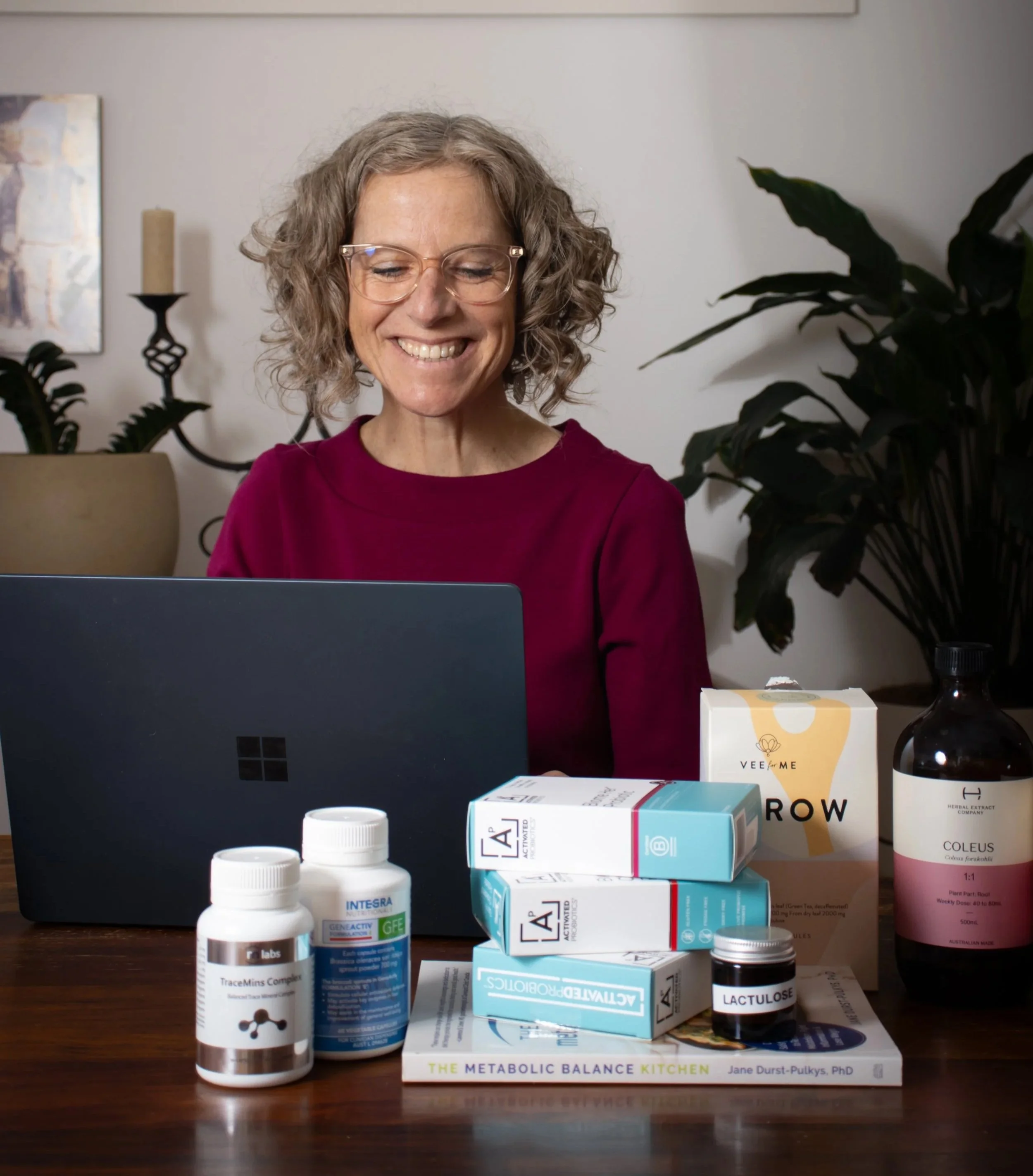 A woman with curly gray hair and glasses smiling at a laptop in a home setting. Various supplement bottles, medication packages, a book titled 'The Metabolic Balance Kitchen,' and a tissue box are on the table in front of her. There are plants and artwork on the white wall in the background.