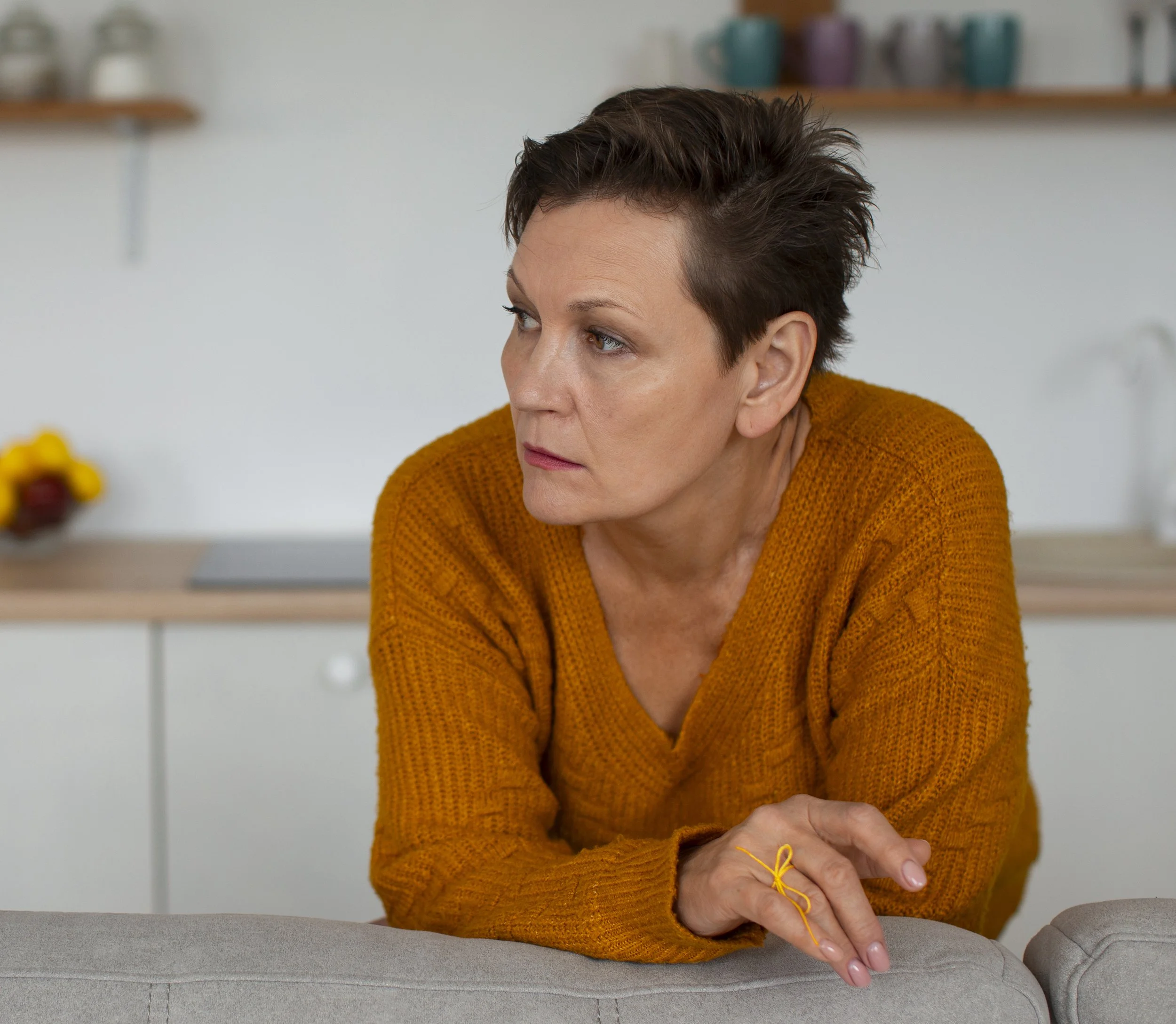 A woman with short, dark hair sits with her arms resting on a cushioned chair, wearing an orange knitted sweater, with a thoughtful expression in a modern kitchen or dining area.