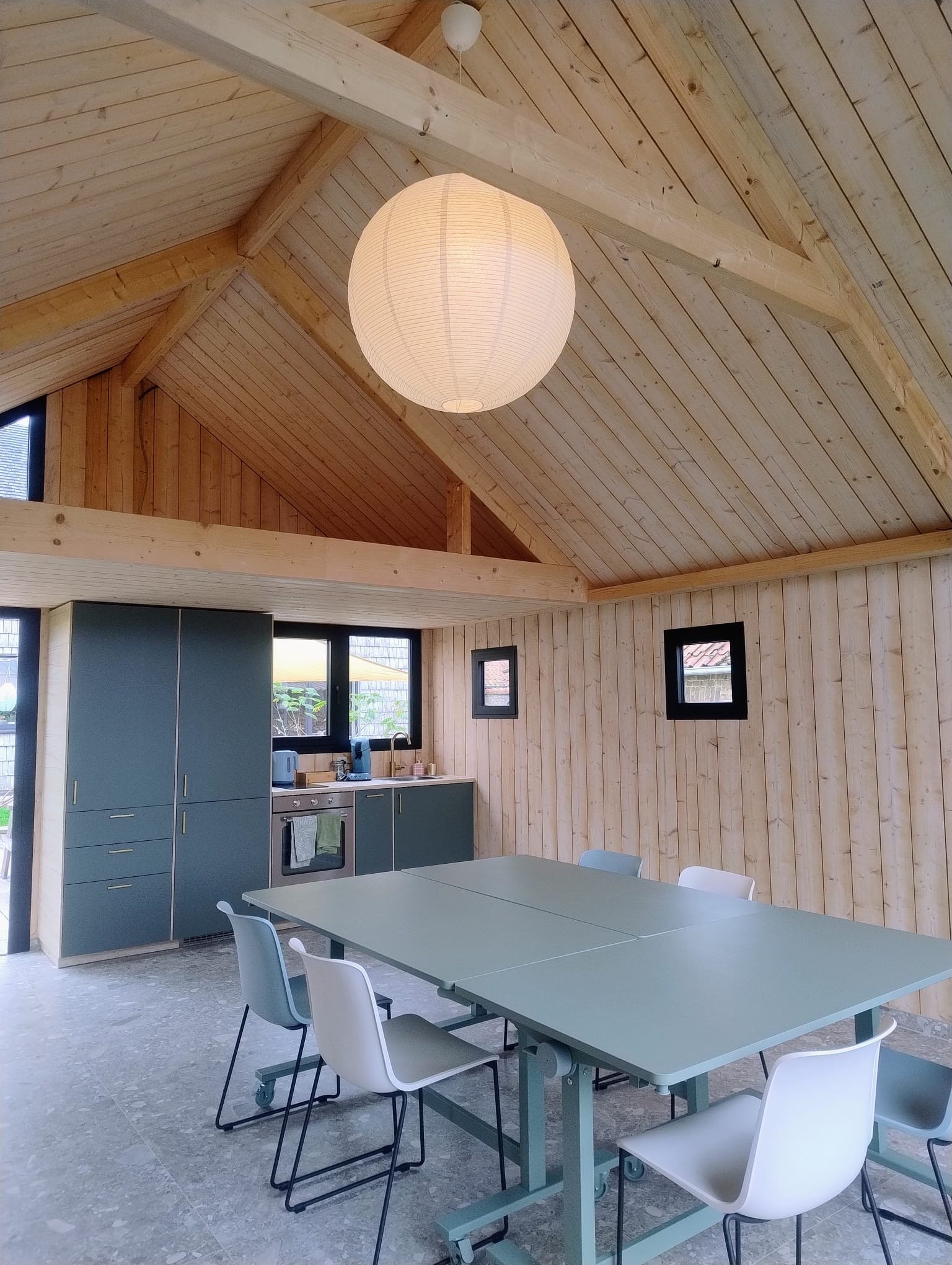 Interieur van een houten huis met een eettafel en stoelen, keukenkasten, ramen en een plafondlamp.