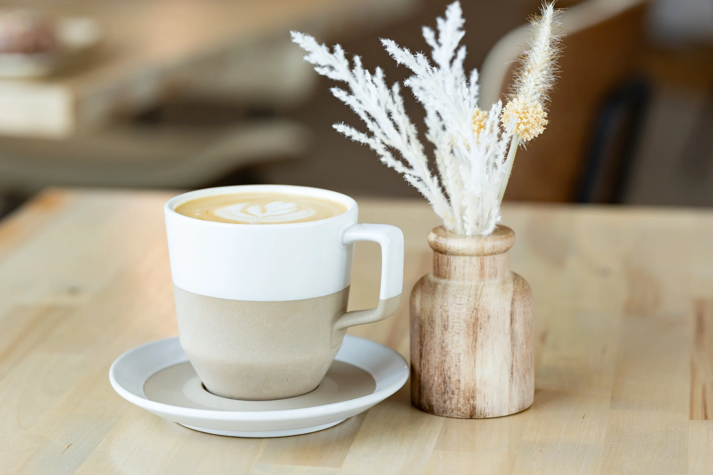 A latte with latte art in a two-tone mug placed on a matching saucer, next to a wooden vase with white dried flowers on a wooden table.