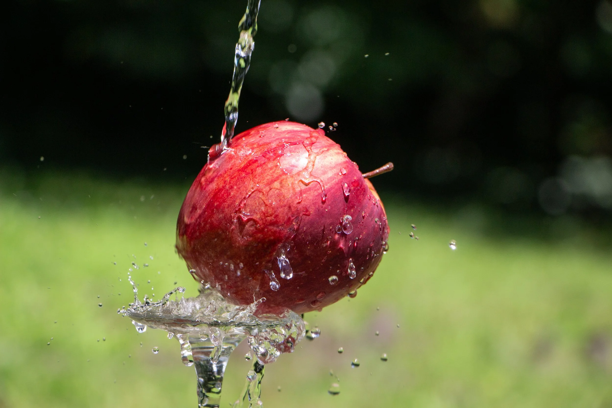 A red apple being washed with water splashing around it against a blurred green background.