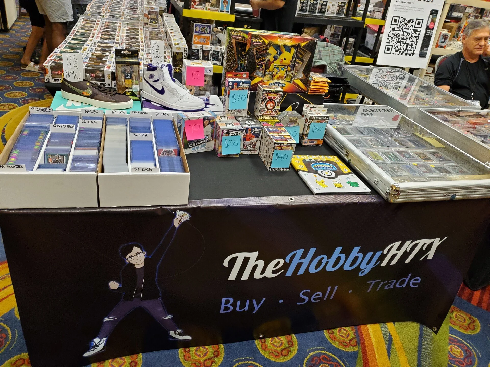 Table at a hobby trade booth with trading card boxes, collectible figures, sneakers, and Pokémon merchandise. Signs indicate prices and various items for sale.