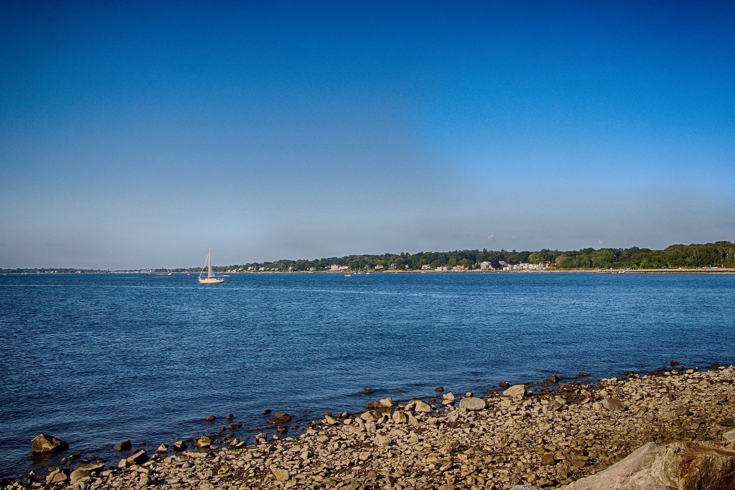 Calm body of water with a sailboat, rocky shoreline in the foreground, and houses along the distant shoreline under a clear blue sky.