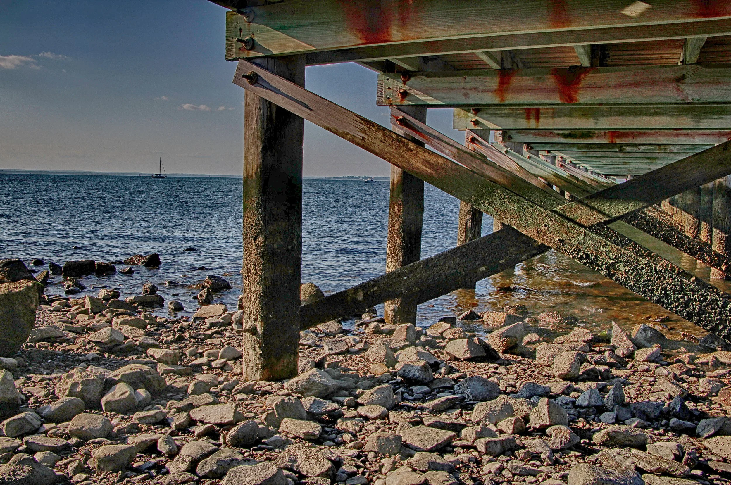 Underwater view of a wooden pier with support beams, overlooking a rocky beach and calm water with a sailboat in the distance.