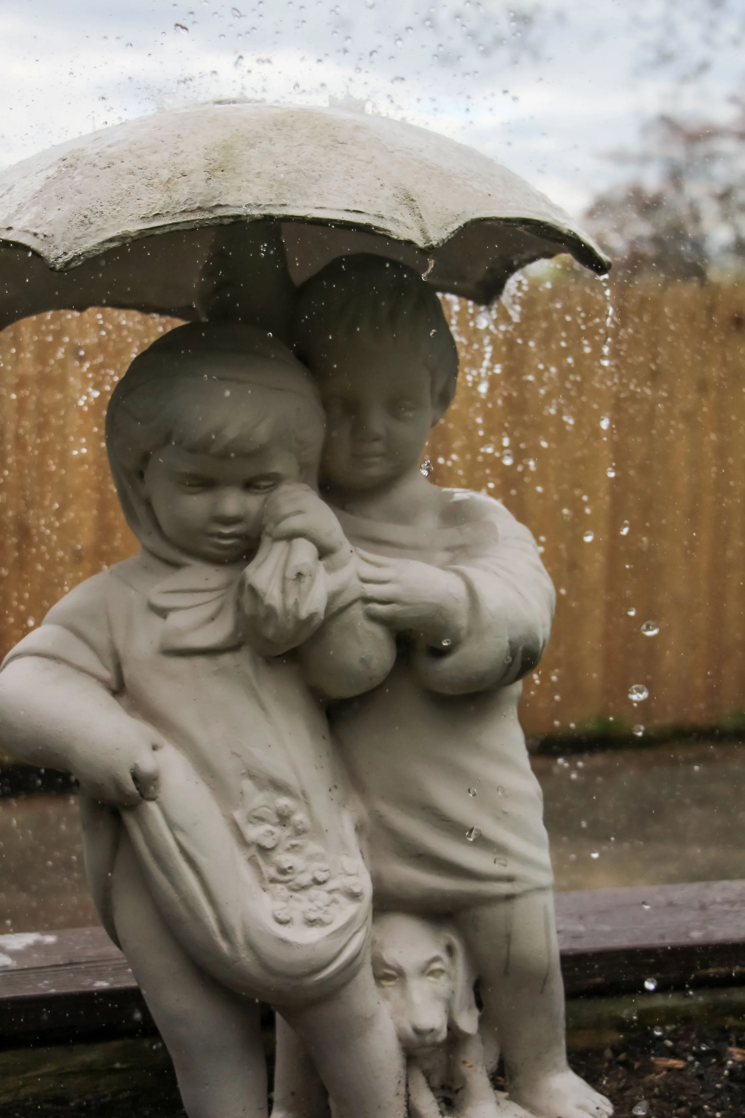 Statue of two children holding an umbrella, with a dog at the base, seen through a rain-covered glass window.