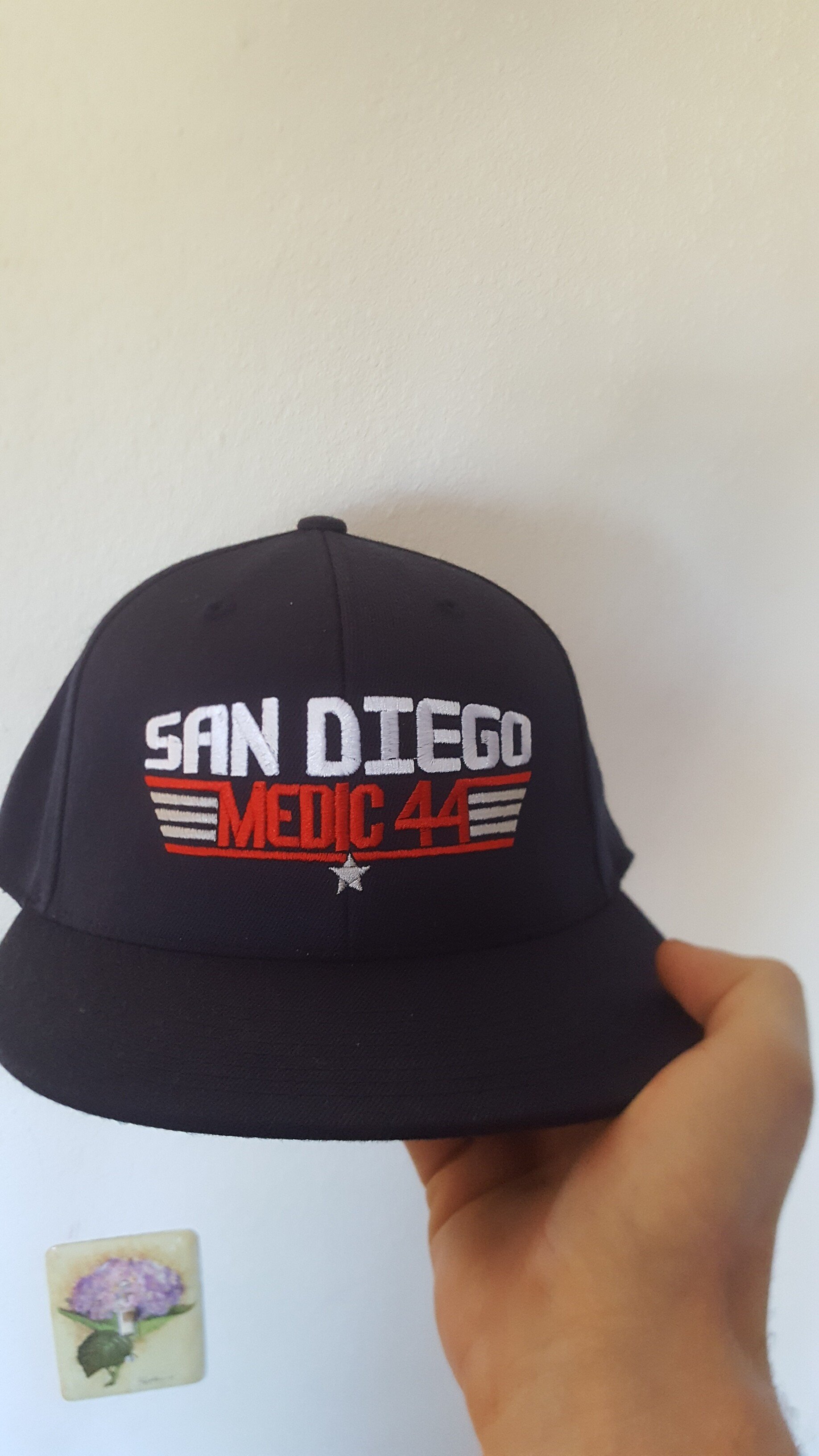 Black baseball cap with embroidered text 'San Diego Medic 44' in white and red, held by a hand against a plain wall with a small flower picture.