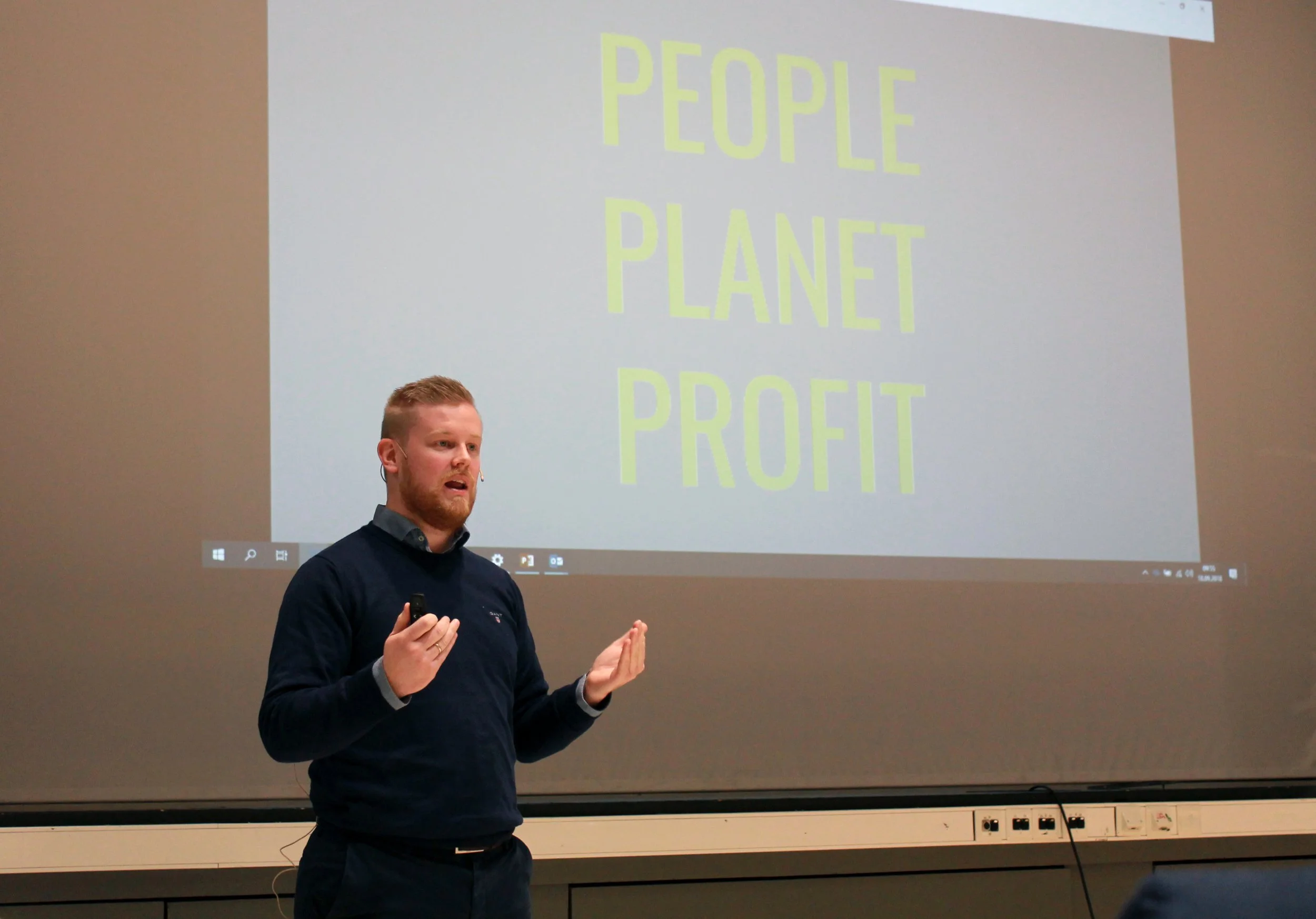 En mann med kort, lyst hår holder en mikrofon og snakker foran en projisert skjerm med store gule bokstaver som sier 'PEOPLE', 'PLANET', og 'PROFIT'.