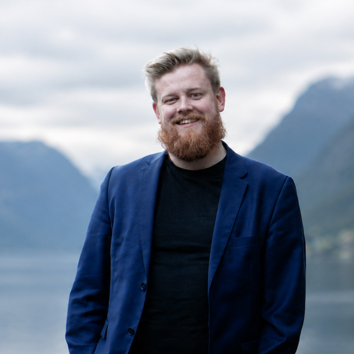 En mann med lyst hår og skjegg smiler, iført en blå blazer og svart t-skjorte, med fjell og vatn i bakgrunnen.