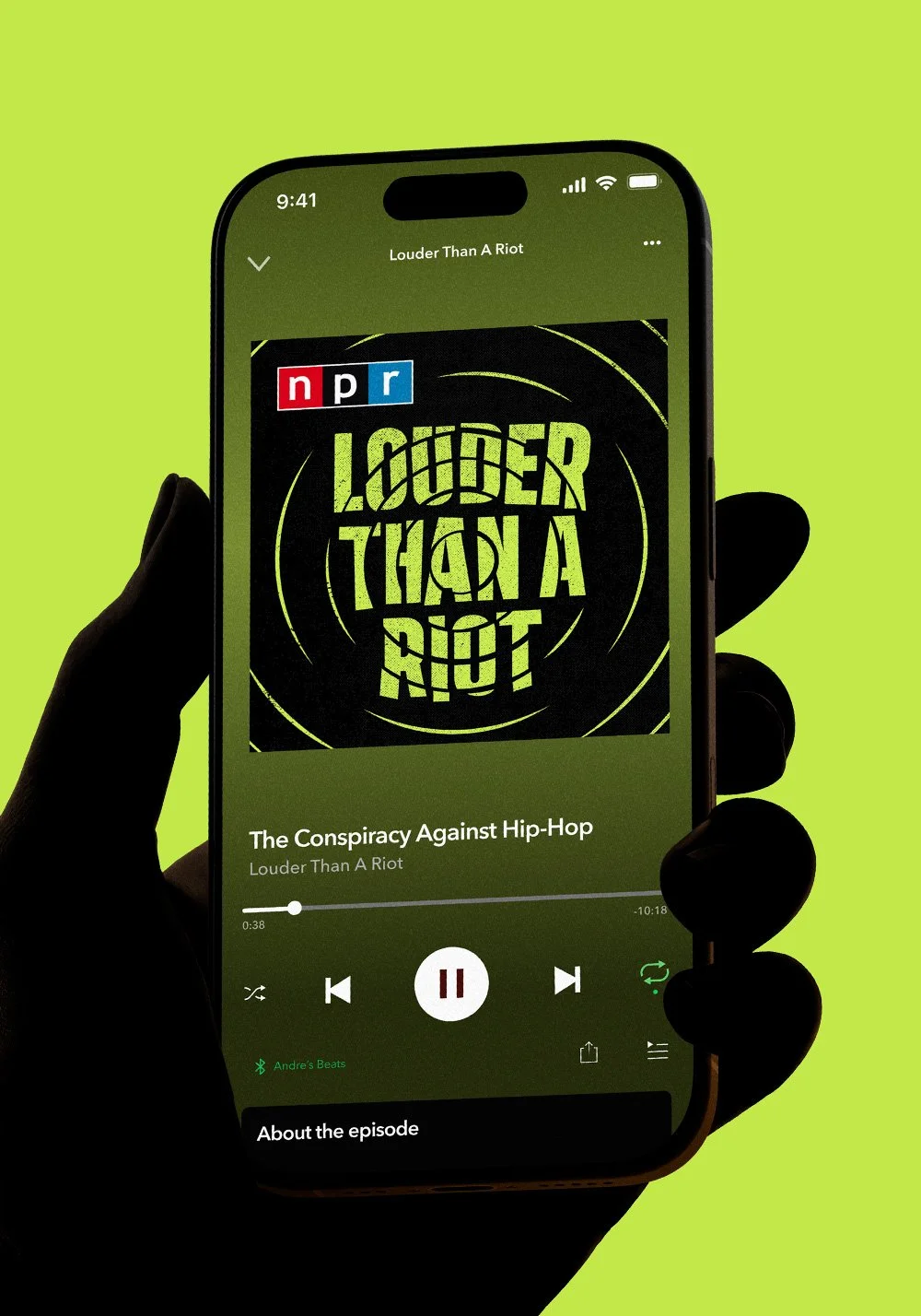 Hand holding a phone displaying the NPR podcast Louder Than A Riot on a green background.