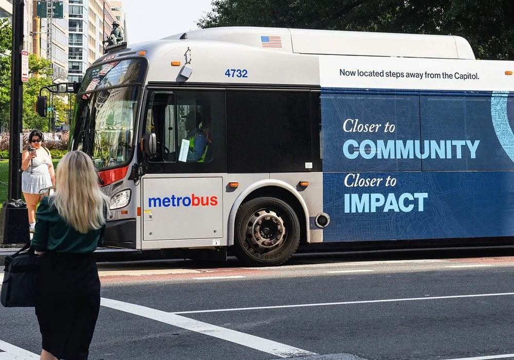 A Metrobus with a 'Closer to Community, Closer to Impact' advertising wrap at a city bus stop, with pedestrians nearby.