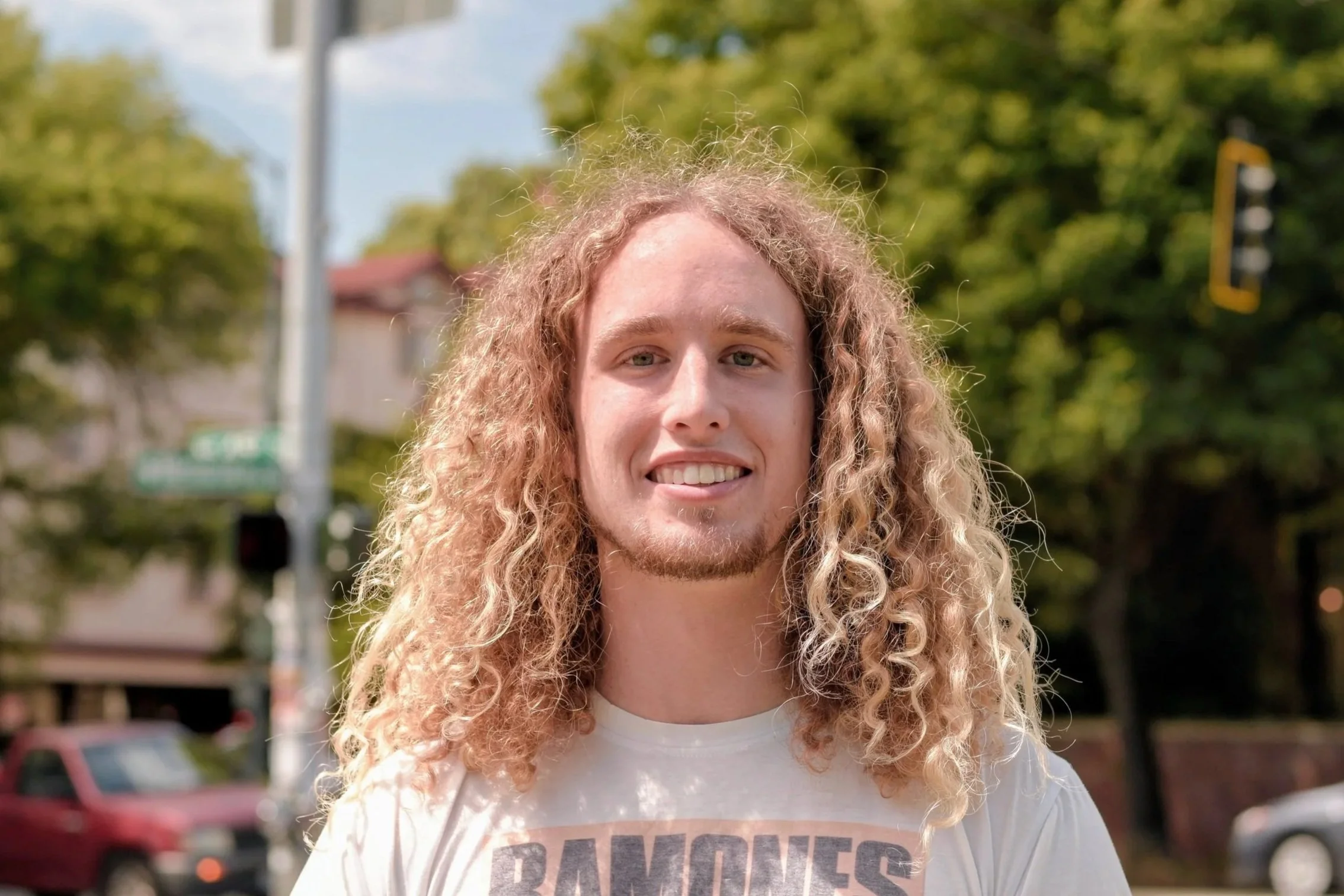 Smiling young man with long curly hair outdoors in a mountainous forest area.