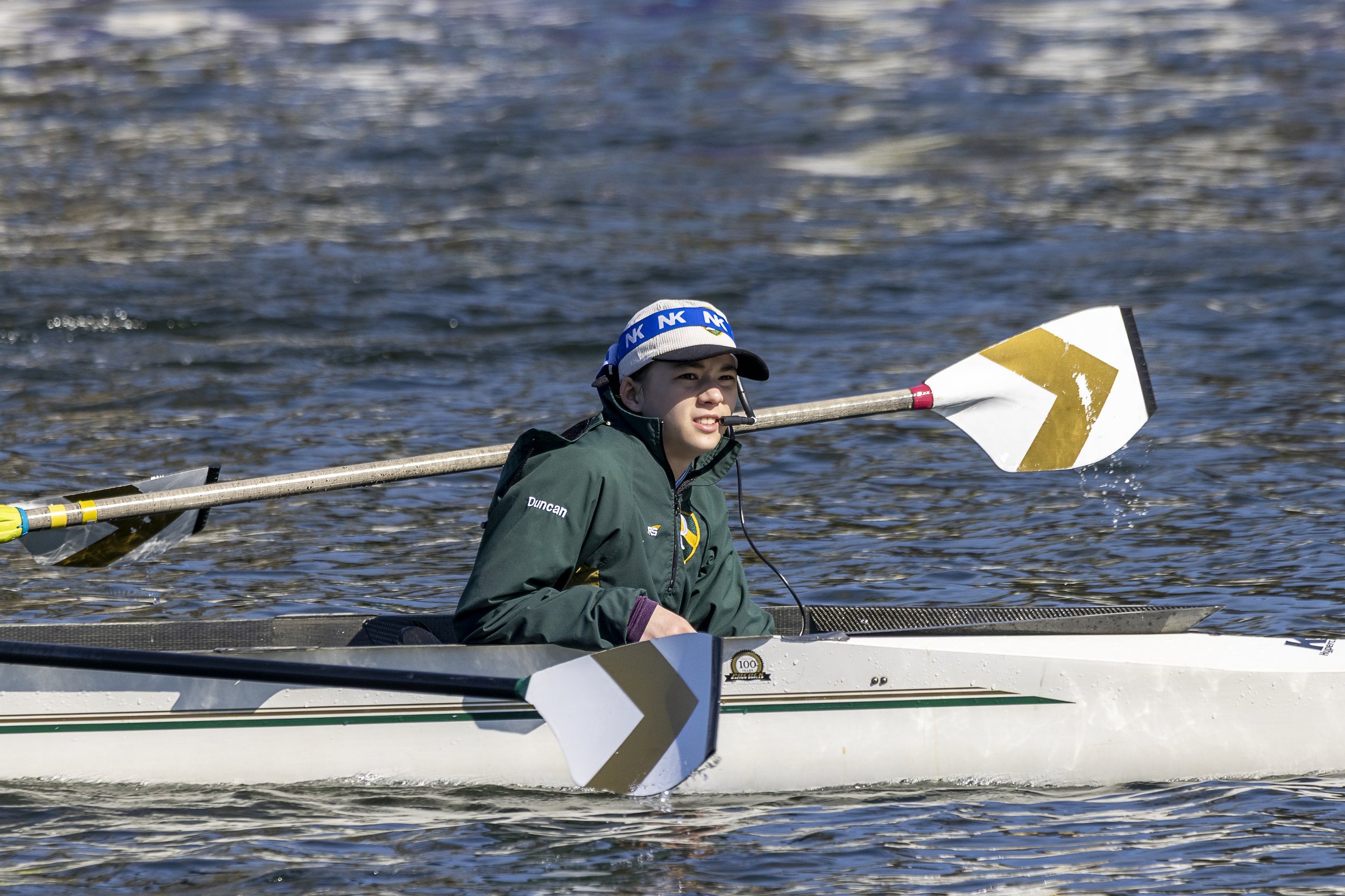 A young male rower with short, light brown hair, wearing red-tinted sports sunglasses, a white and green rowing uniform, and carrying a racing shell on his shoulder during a rowing event.