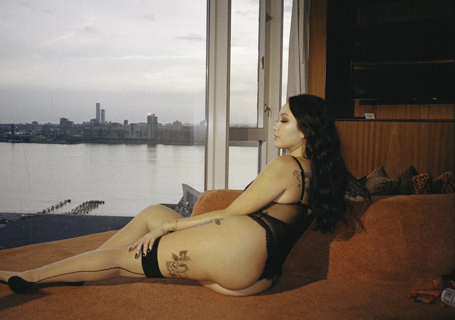 A woman with long black hair and tattoos in black lingerie, sitting on an orange sofa, facing a large window overlooking a city skyline and water during sunset or evening.