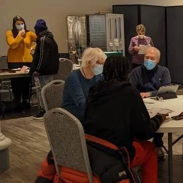 People sitting at tables and wearing face masks in a conference room, with some individuals standing in the background.