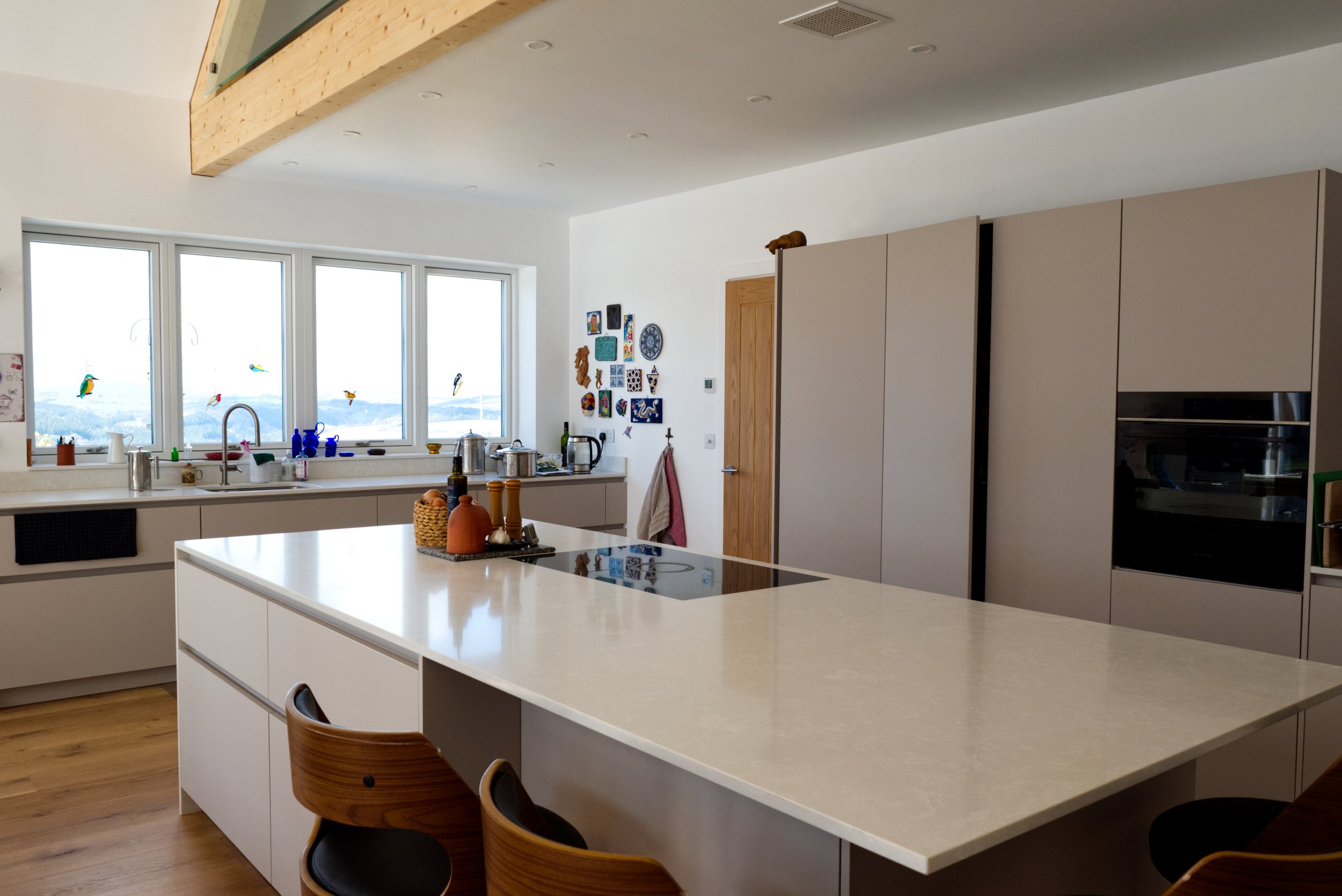 Modern kitchen with a white island, wooden chairs, large windows, and minimalist cabinetry.