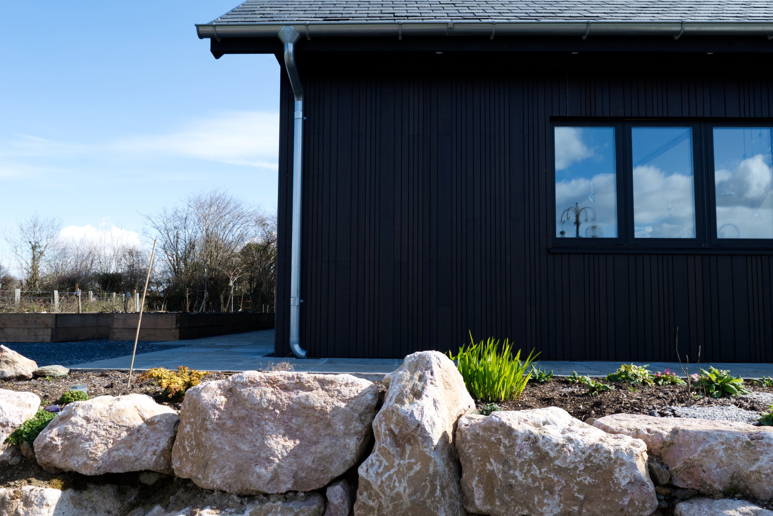Exterior of a dark-colored modern building with large windows, a downspout, and a shingle roof. In the foreground, there's a garden bed with rocks and plants. The background shows a clear sky and some trees.