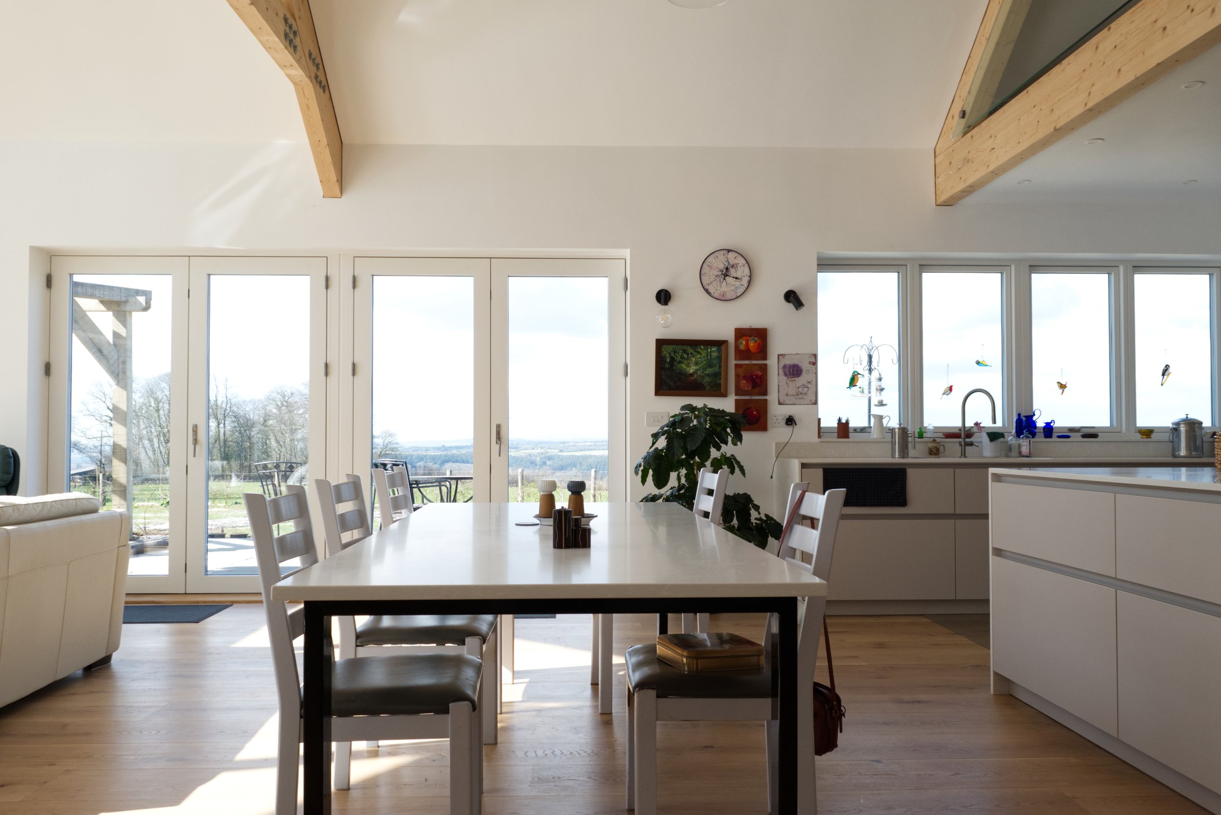 Modern kitchen and dining area with a large table and chairs, decorative wall clock, windows offering a view of the outdoors, a countertop with dishes and a plant, and wooden beam ceiling accents.