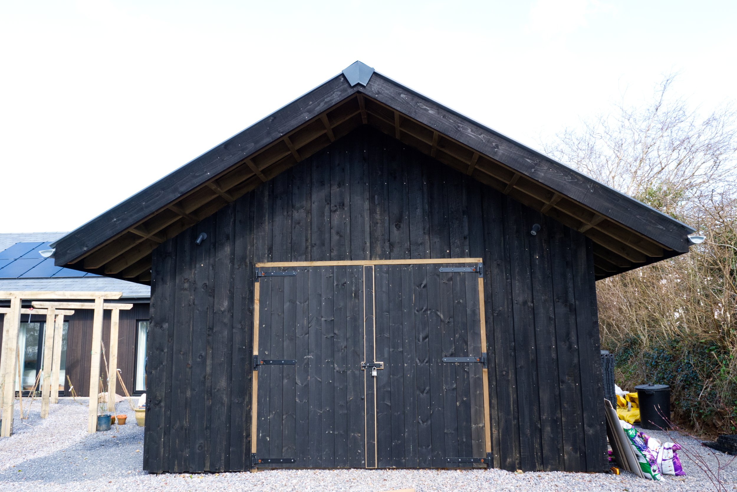 Dark wooden barn with large double doors, gravel path, and some garden supplies on the side.