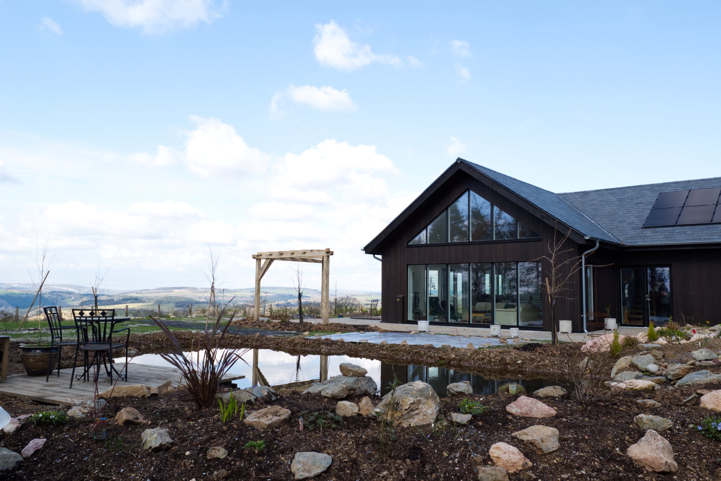 Modern house with large windows, patio, and garden pond in a rural setting.