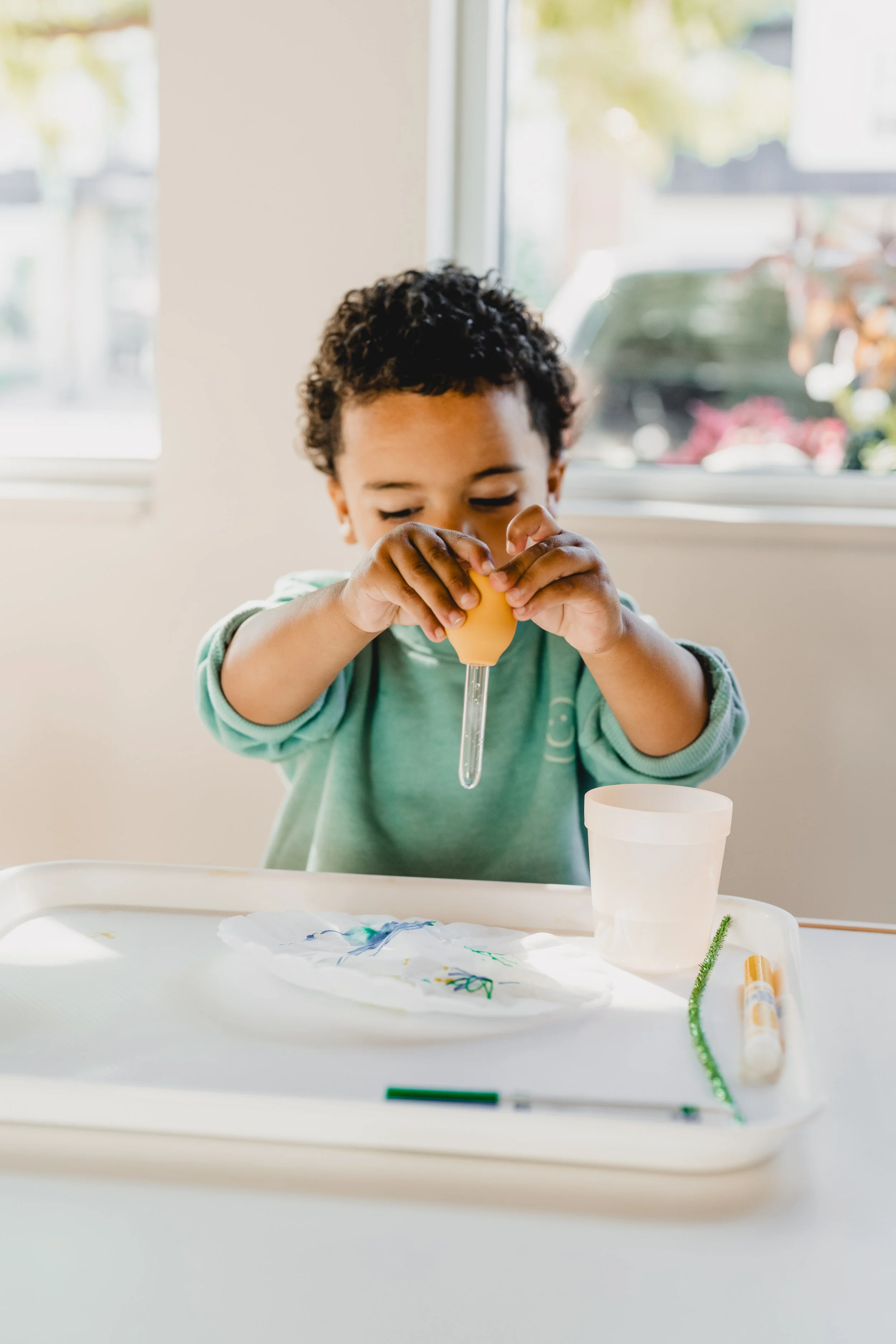 Young child with a white bow in her hair holding and decorating colorful balls on a paper plate at a table.