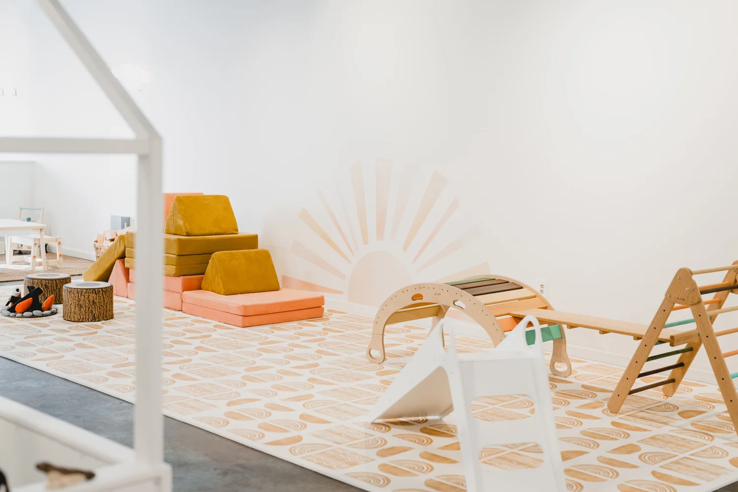 The Play Group's bright indoor play space with a sunburst mural, soft seating, and wooden climbing toys