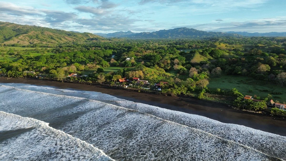 Aerial view of a coastal landscape with a black sand beach, rolling waves, lush green hills, scattered houses, and distant mountains under a partly cloudy sky.