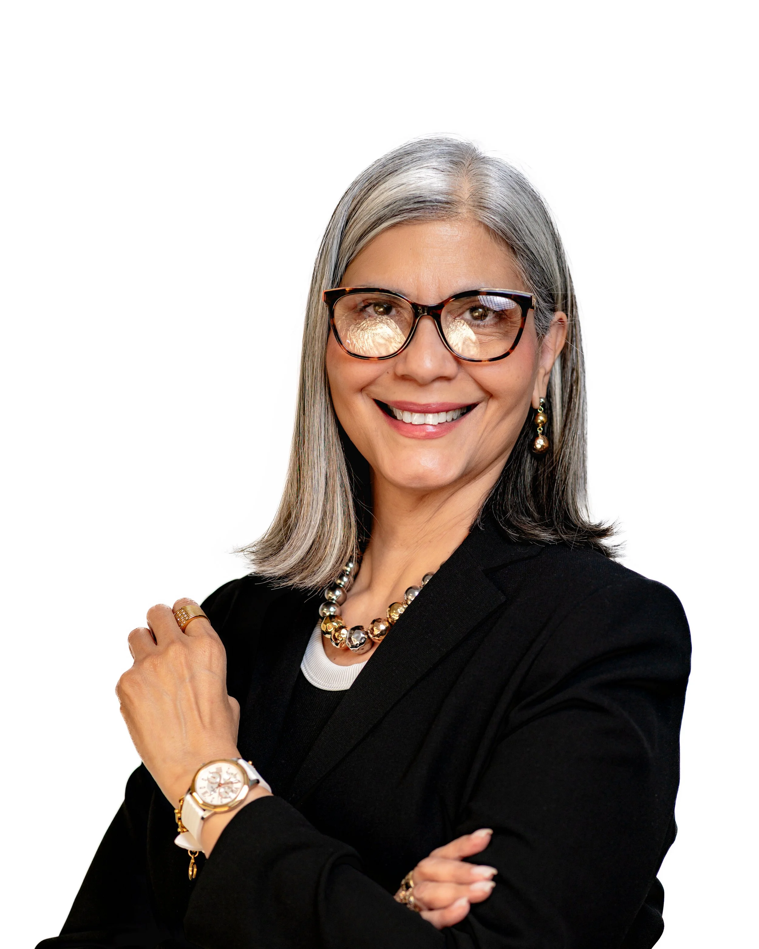 A smiling woman with shoulder-length gray hair sitting on patterned furniture in a well-lit room, wearing a white blazer with black stripes on the sleeves, a black turtleneck, a pearl necklace, and a bracelet.