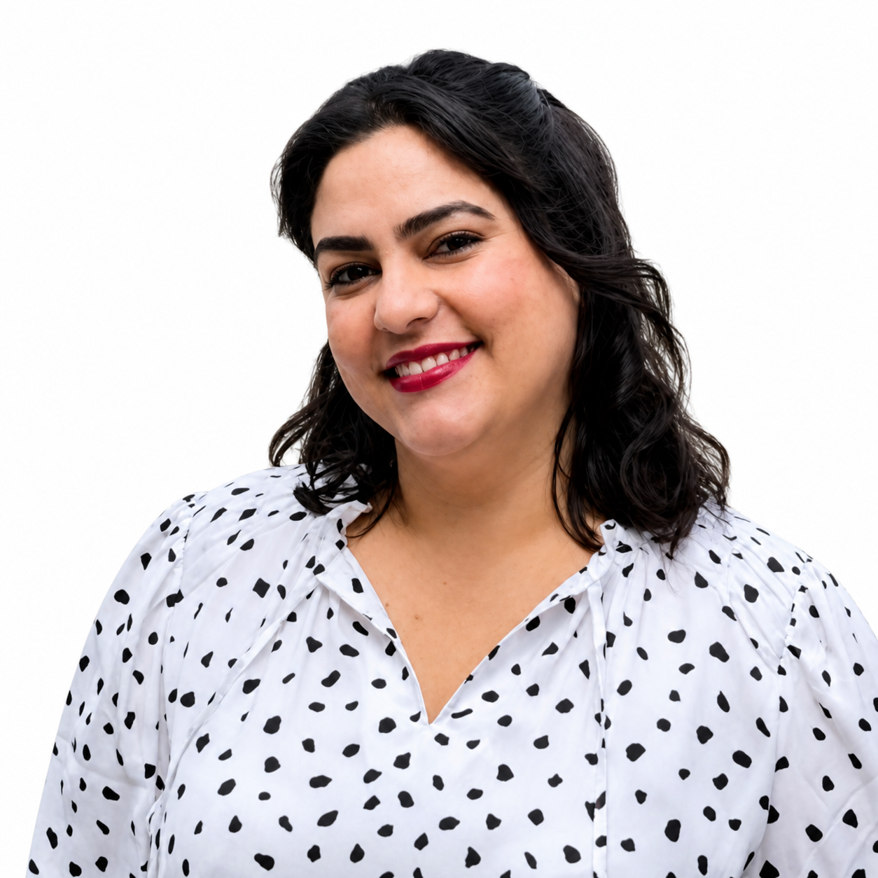 A woman with dark hair and red lipstick smiling outdoors, wearing a white blouse with black polka dots, in a park with trees and grass in the background.