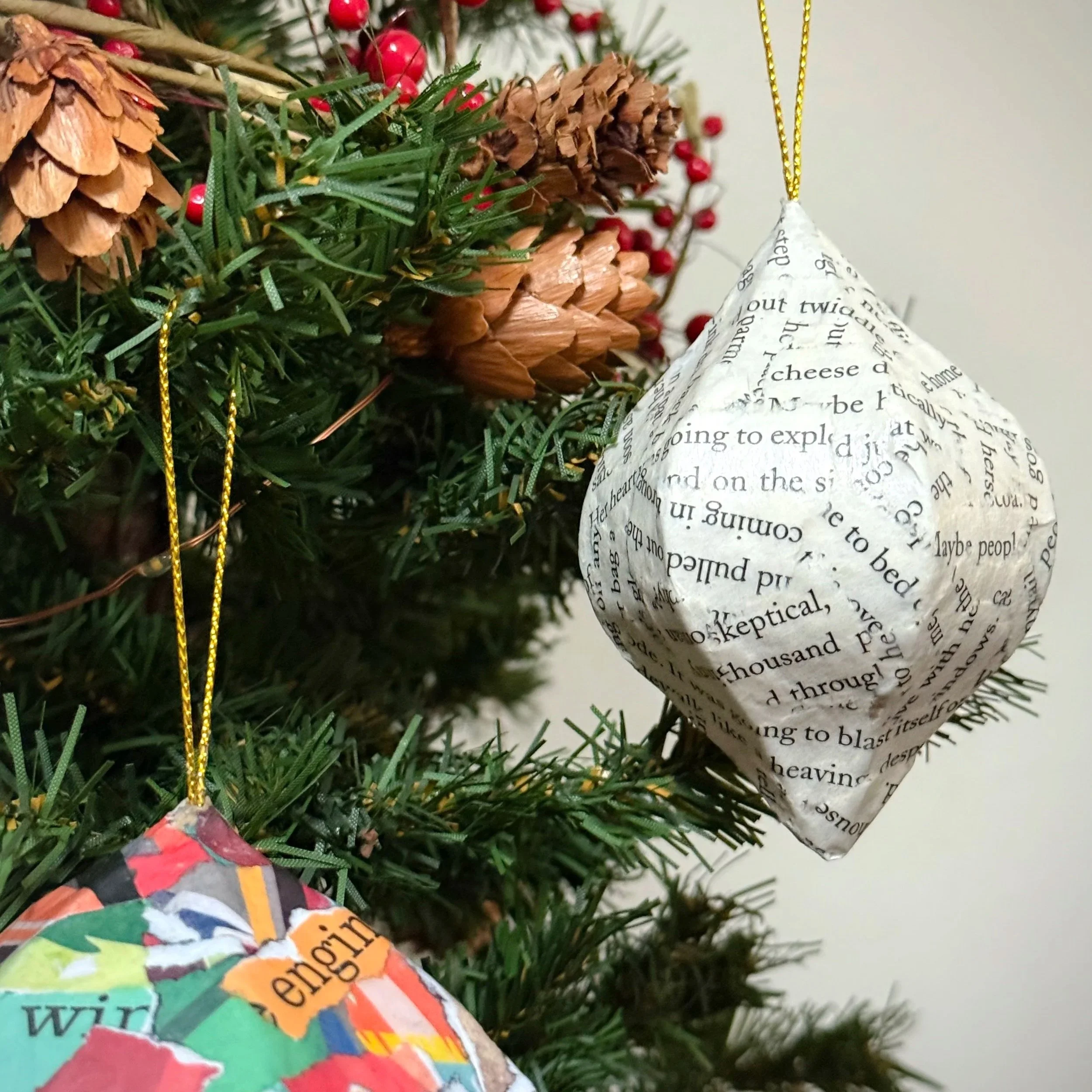 Christmas tree with two handmade book page ornaments, one bright and colorful and one in muted black-and-white text, hanging among pinecones and red berries.