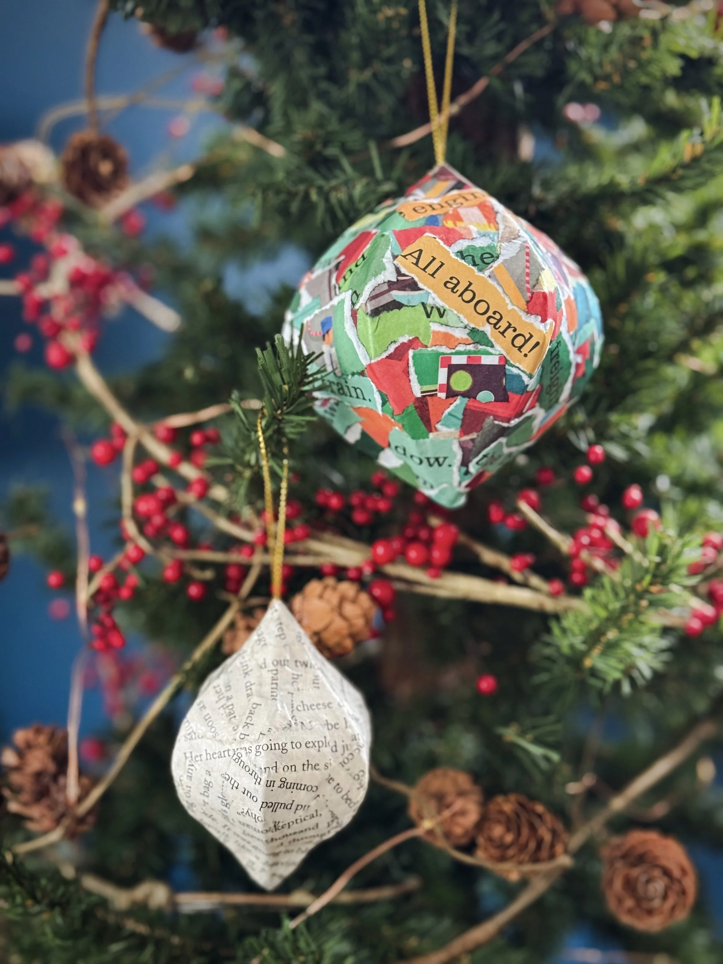 Finished book page Christmas ornaments hanging on a decorated tree: one colorful train-themed ornament from "On The Train" and one neutral text-only ornament made from a novel.