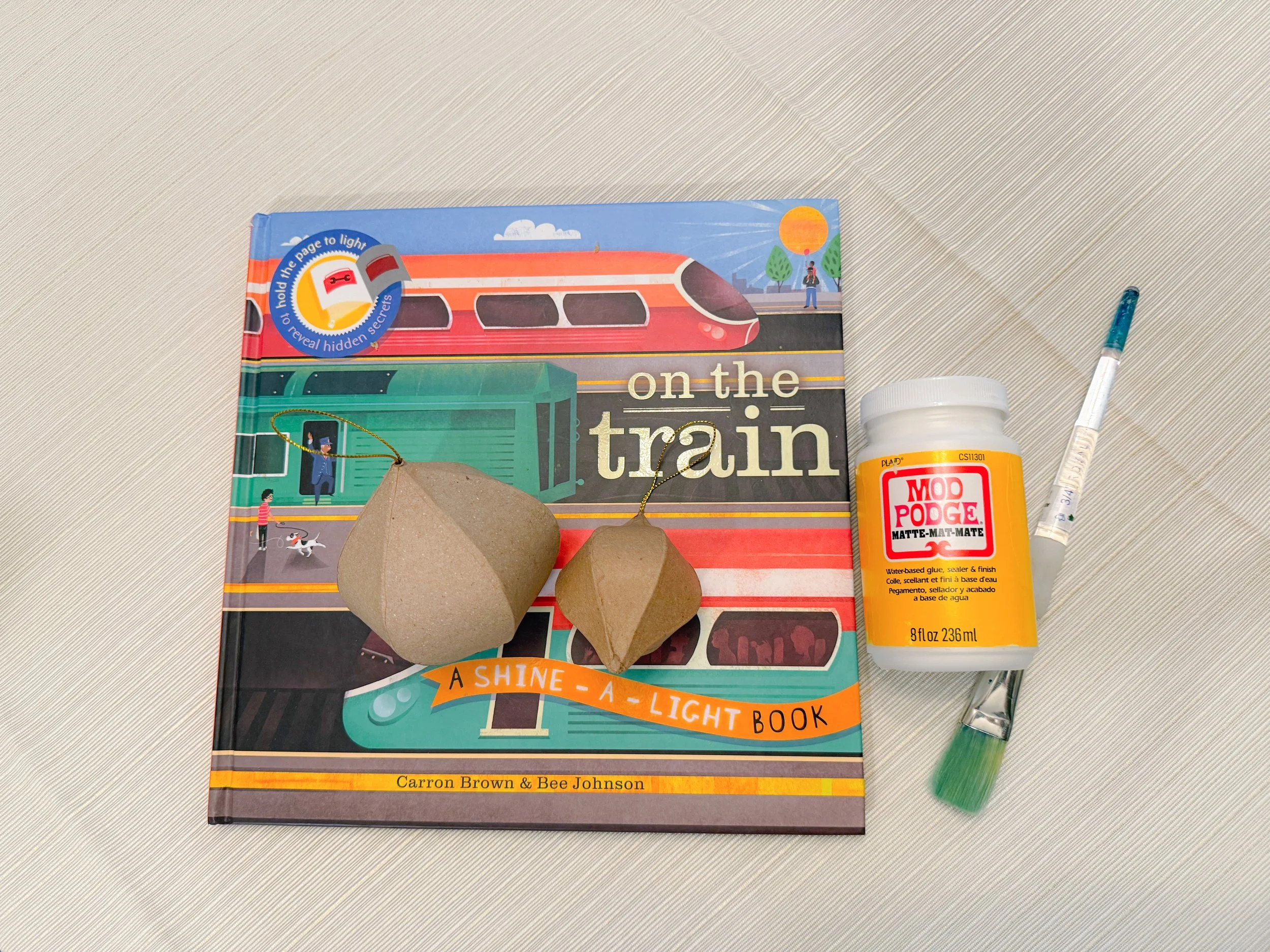 Two brown paper craft ornaments sit on top of a colorful picture book on a table, ready for a family craft activity. Next to the picture book is a paintbrush and a bottle of Mod Podge.