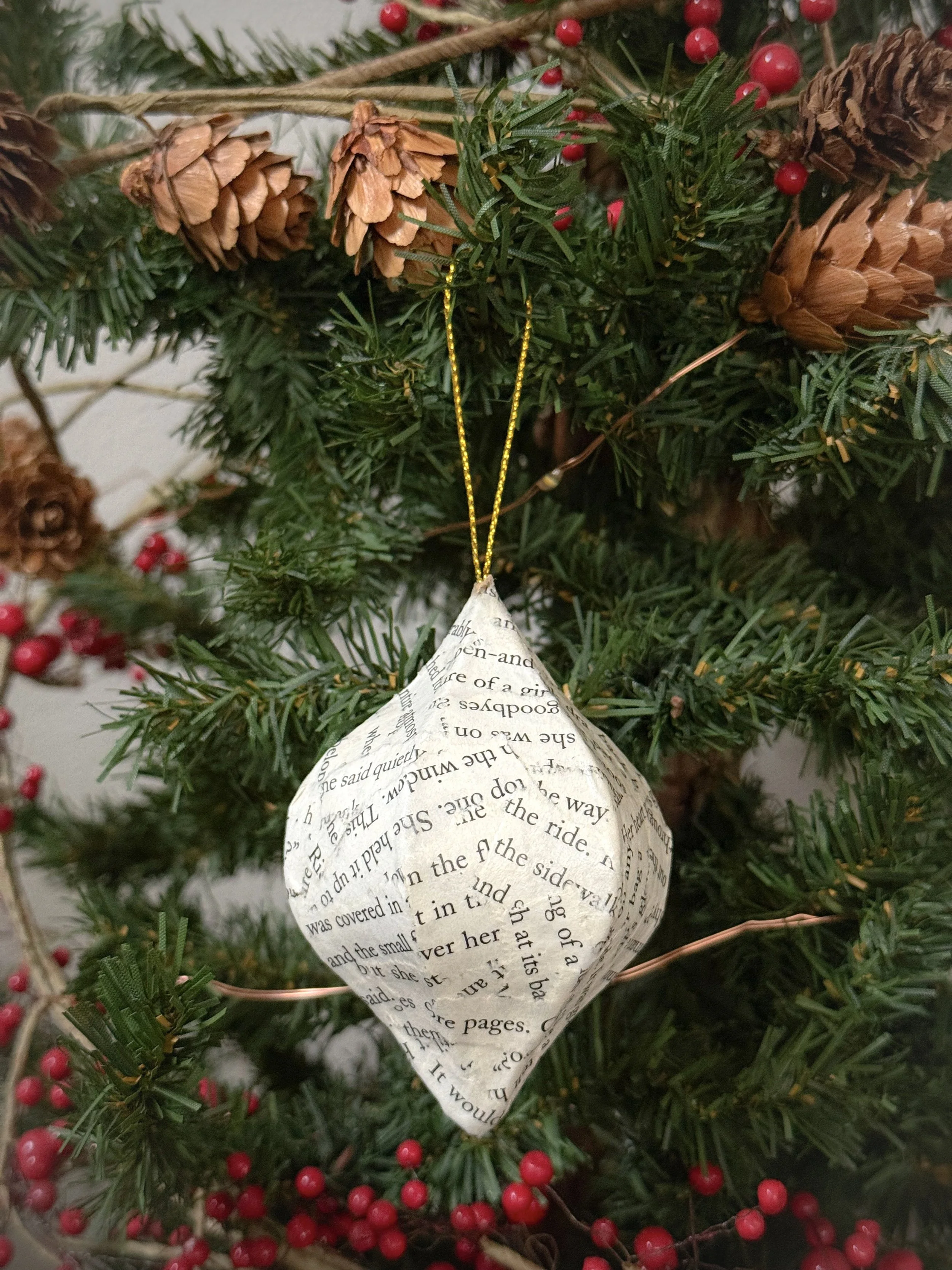 Close-up of the neutral text-only book page ornament made from decoupaged paper strips, hanging on a tree with pine cones and red berries.