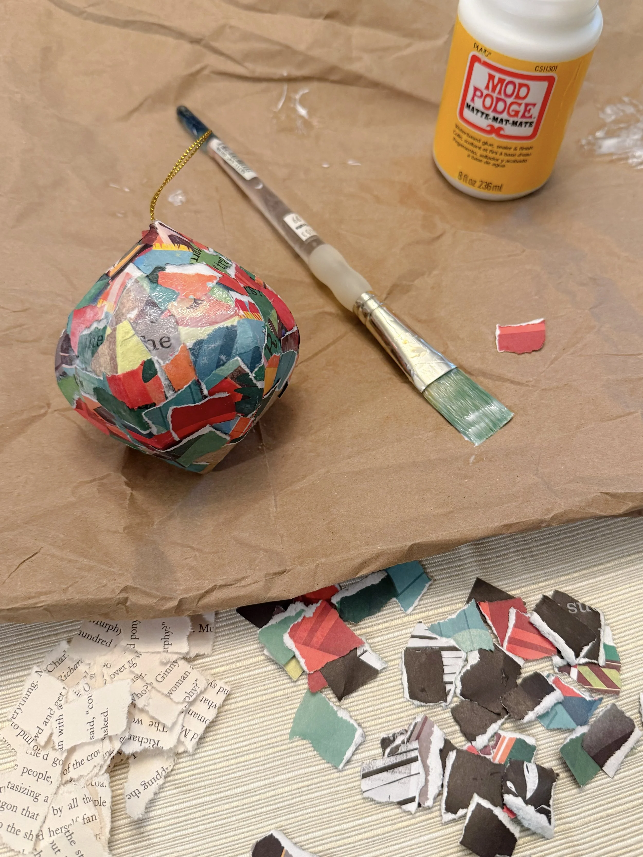 In-progress DIY book page ornament covered in colorful torn book page pieces, surrounded by more paper scraps, a paintbrush, and a jar of Mod Podge.