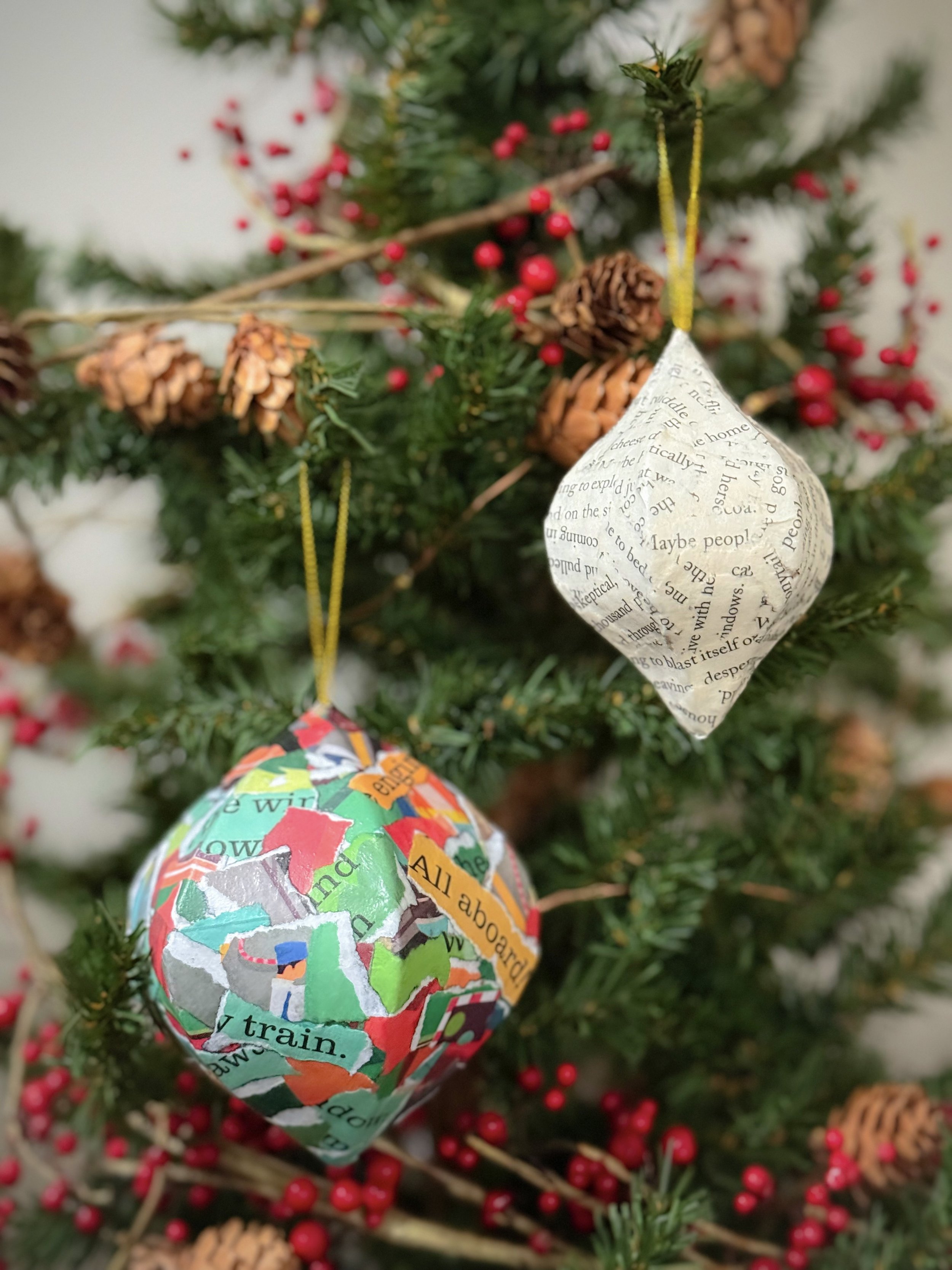 Family Christmas craft ornaments made from picture books and novels are hanging in a tree adorned with pinecones and red berries.