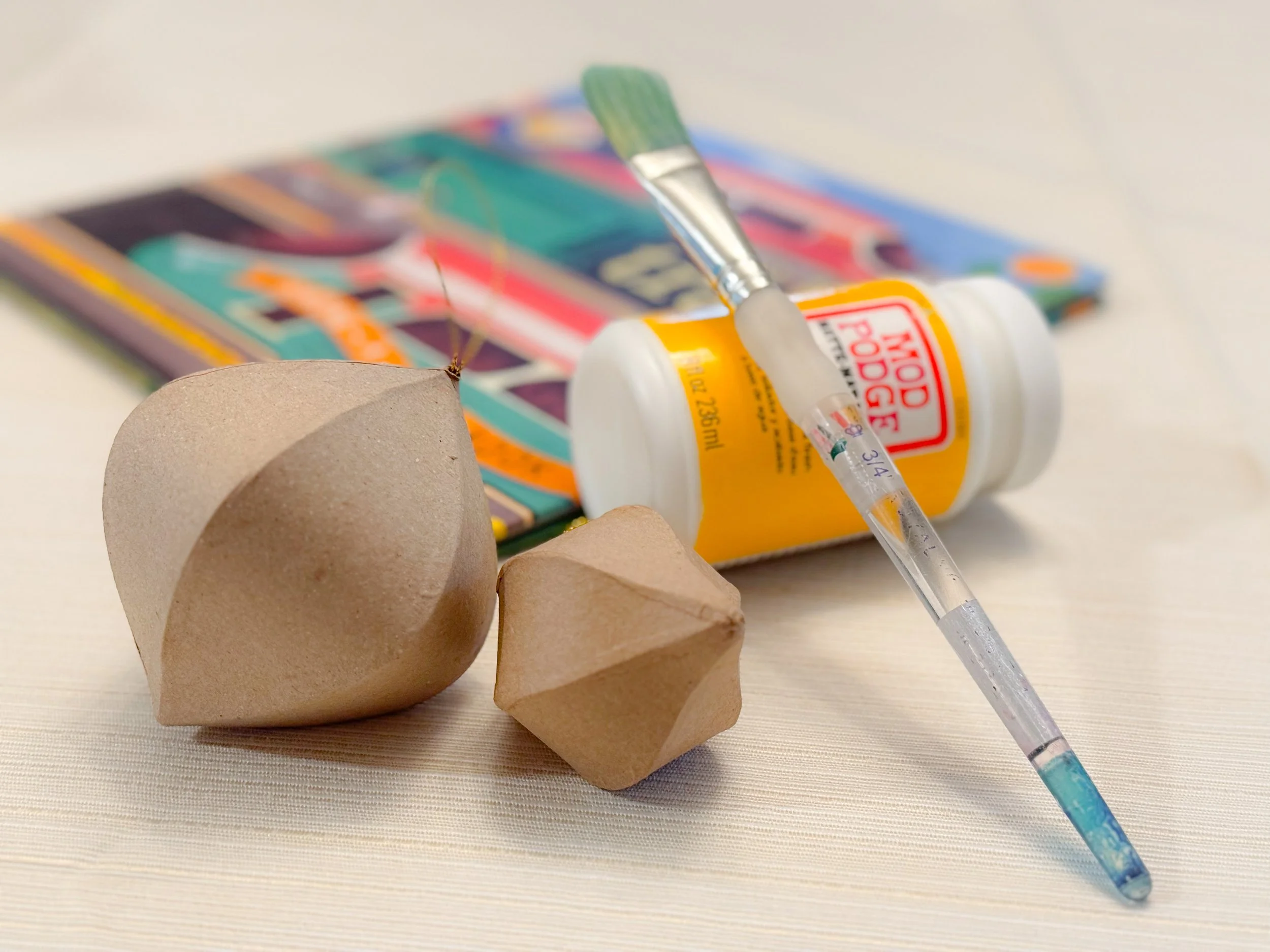 Supplies for DIY book page Christmas ornaments: two plain brown paper ornaments, a jar of Mod Podge, a paintbrush, and a colorful picture book in the background on a light table.
