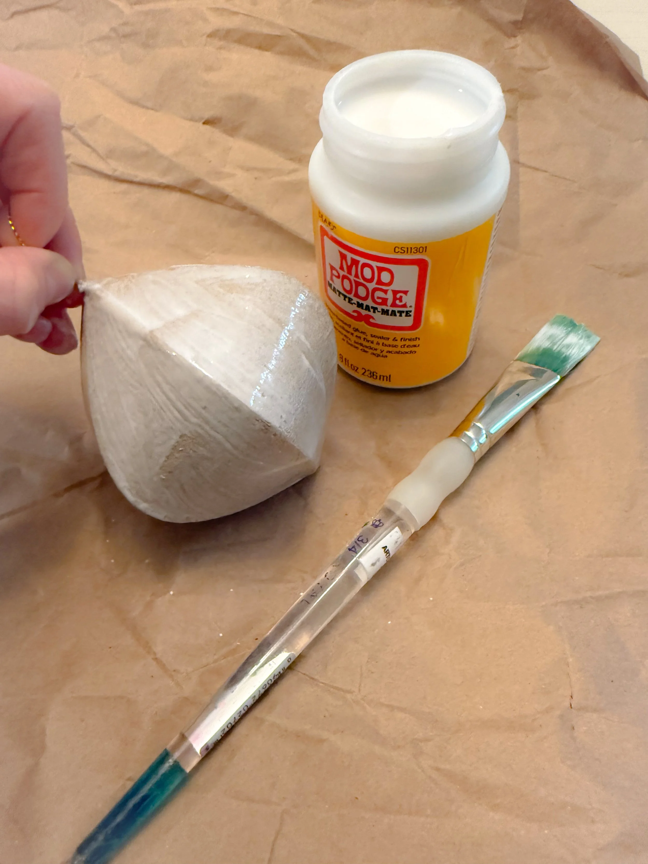 Brown paper ornament being coated with Mod Podge, with an open jar of glue and a paintbrush resting on kraft paper underneath.