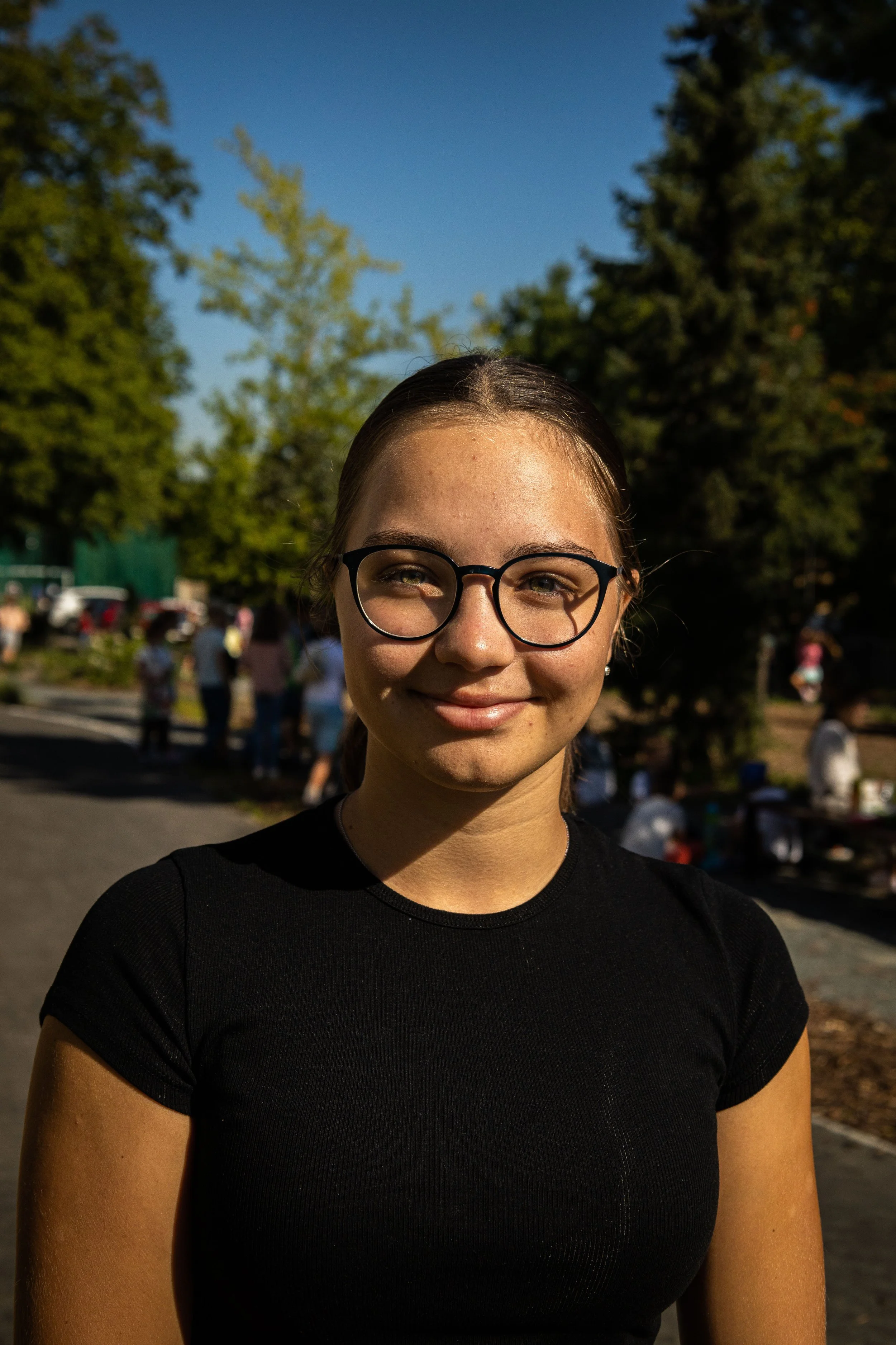A young woman with glasses and a black shirt smiling outdoors with trees and people in the background.