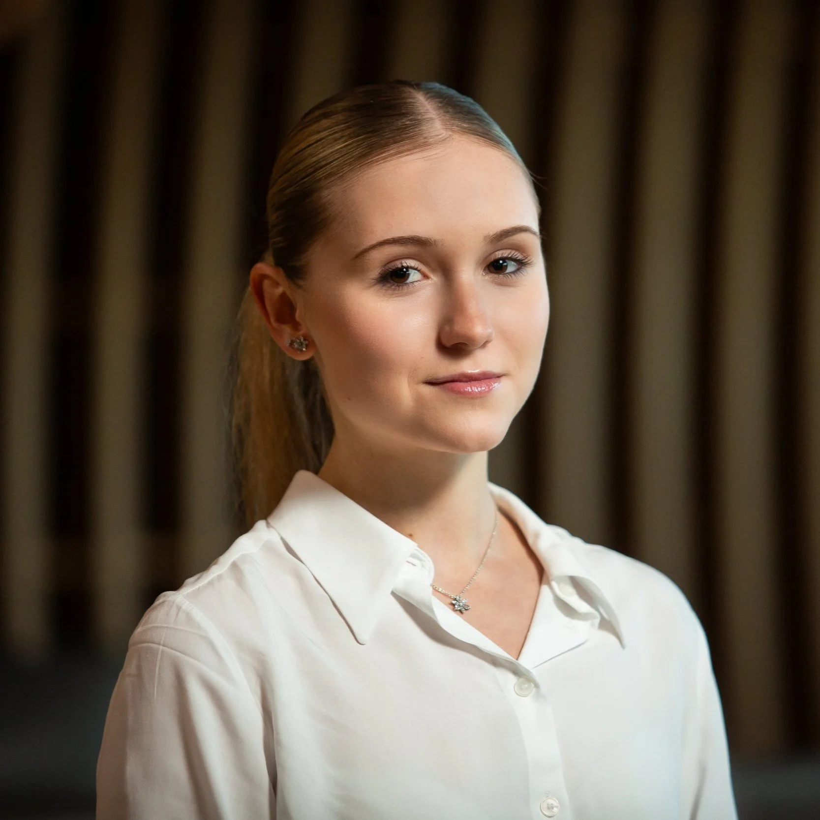A young woman with light skin, blonde hair tied back, wearing a white blouse, silver earrings, and a necklace, standing indoors with a neutral background.