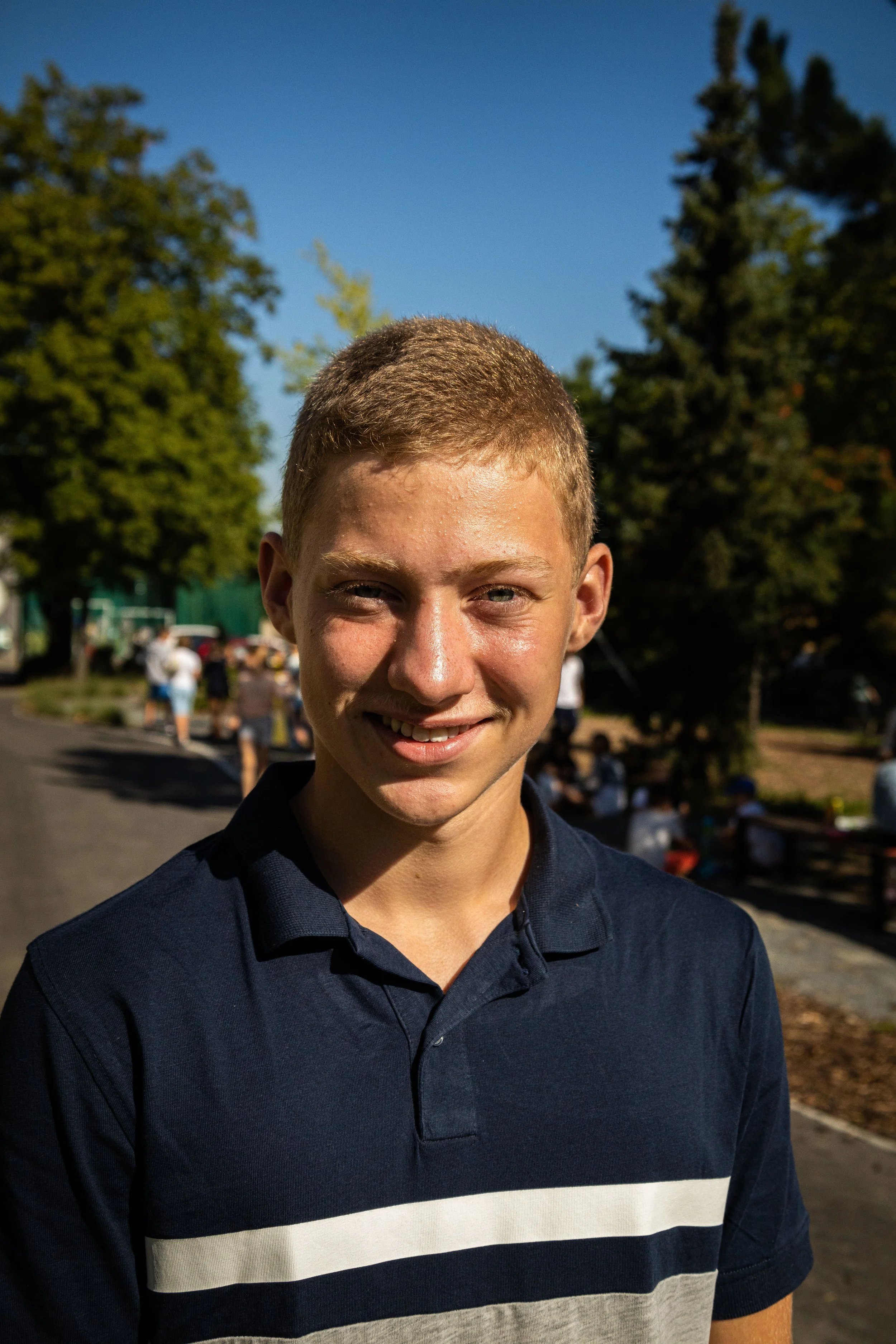 Young smiling boy with short blond hair in a navy blue polo shirt outdoors on sunny day, with trees and people in the background.