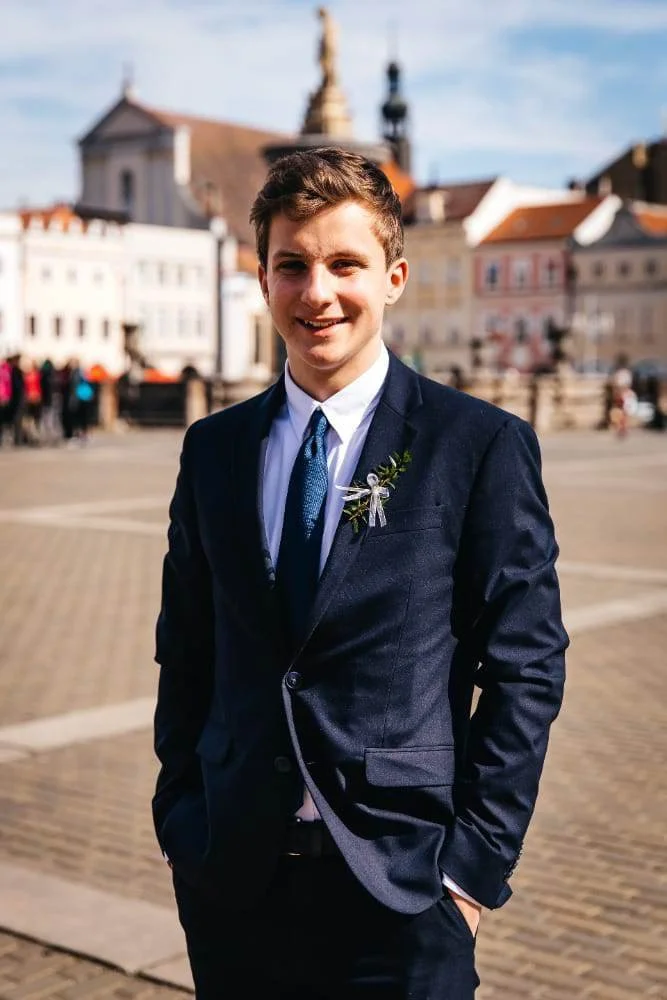 A young man in a dark suit and tie standing outdoors in a city square, smiling with his hands in his pockets, with historic buildings and a church in the background on a sunny day.