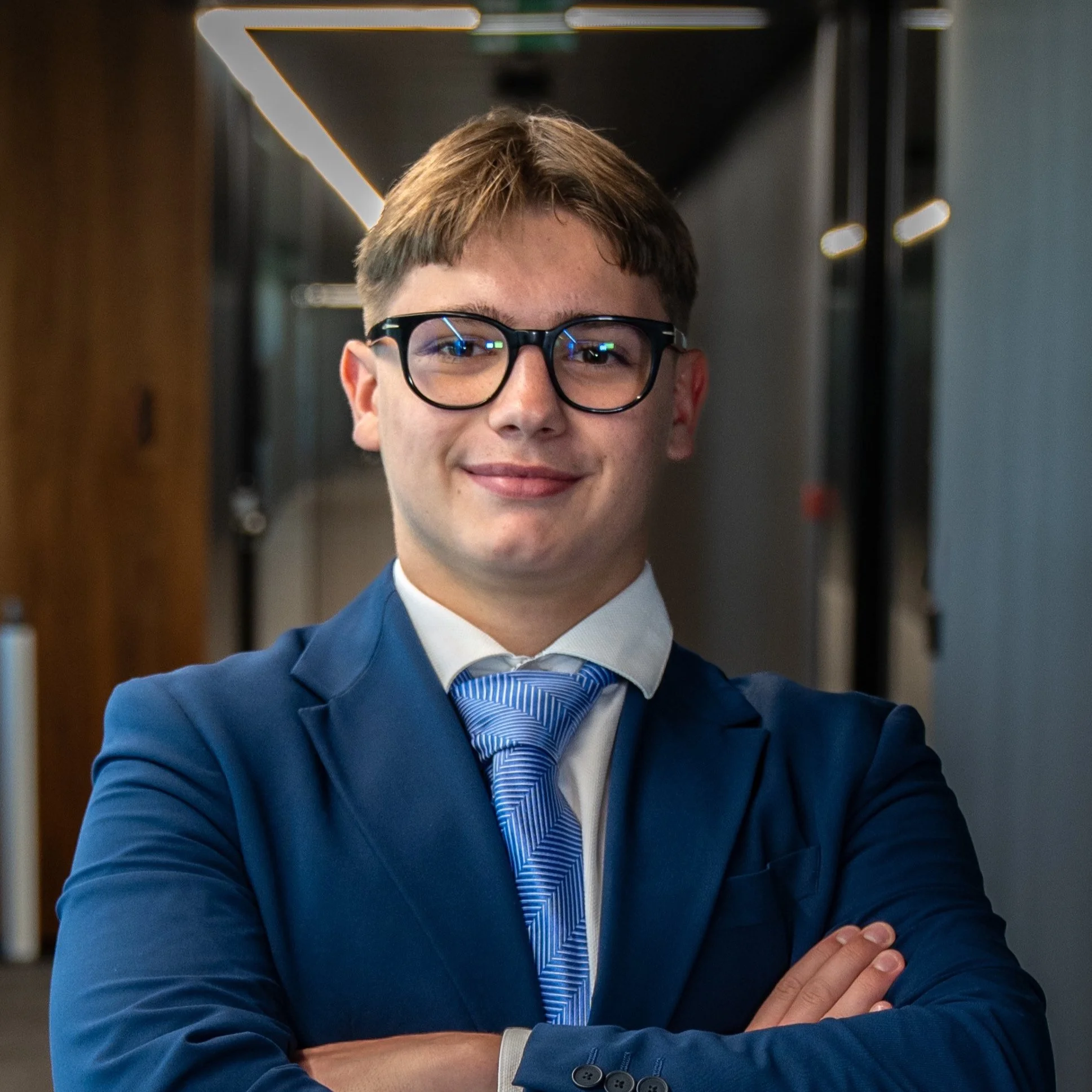 A young man in a navy blue suit and striped blue tie, wearing glasses, smiling with arms crossed in a modern professional setting.