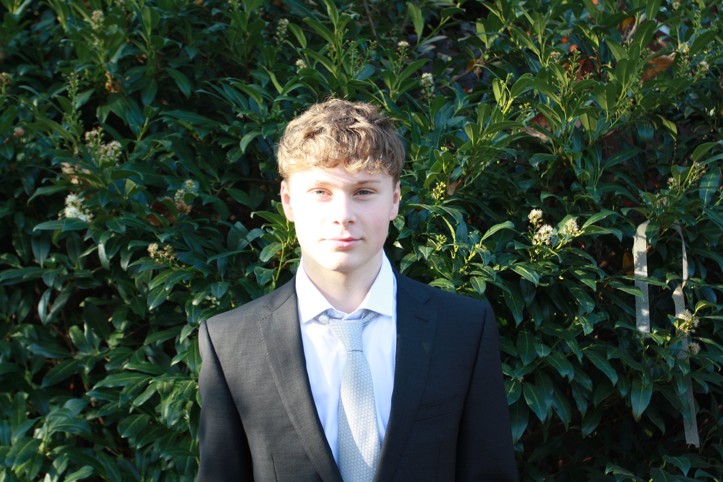 A young man in a black suit, white shirt, and light gray tie standing outdoors in front of green leafy bushes.