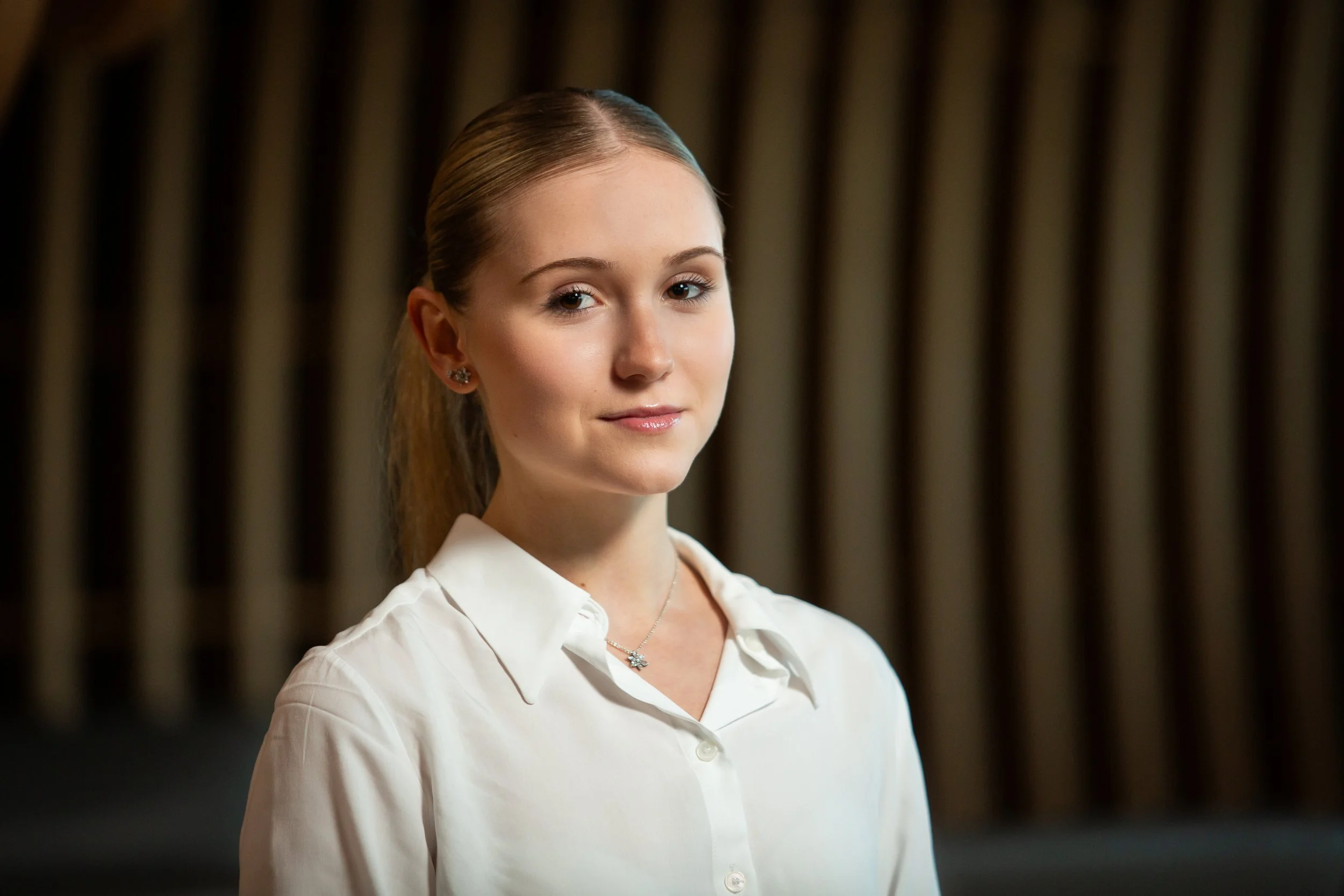 A young woman with light brown hair in a ponytail, wearing a white blouse and small earrings, standing against a blurred background with vertical brown panels.