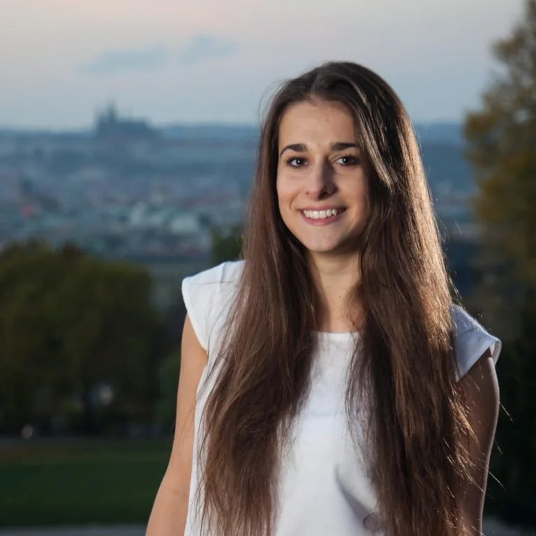 A young woman with long brown hair, smiling, wearing a white sleeveless top, outdoors with blurred trees and a cityscape in the background.