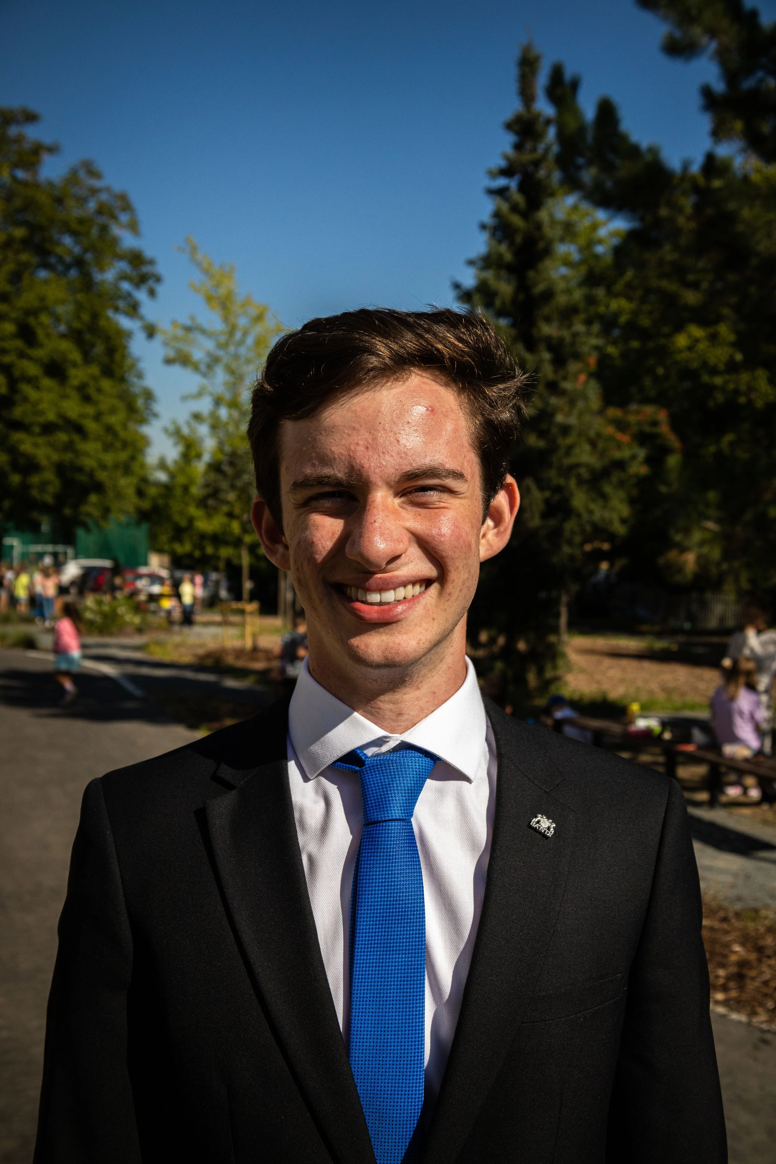 Young man in a black suit, white shirt, and blue tie smiling outdoors on a sunny day with trees and a park in the background.