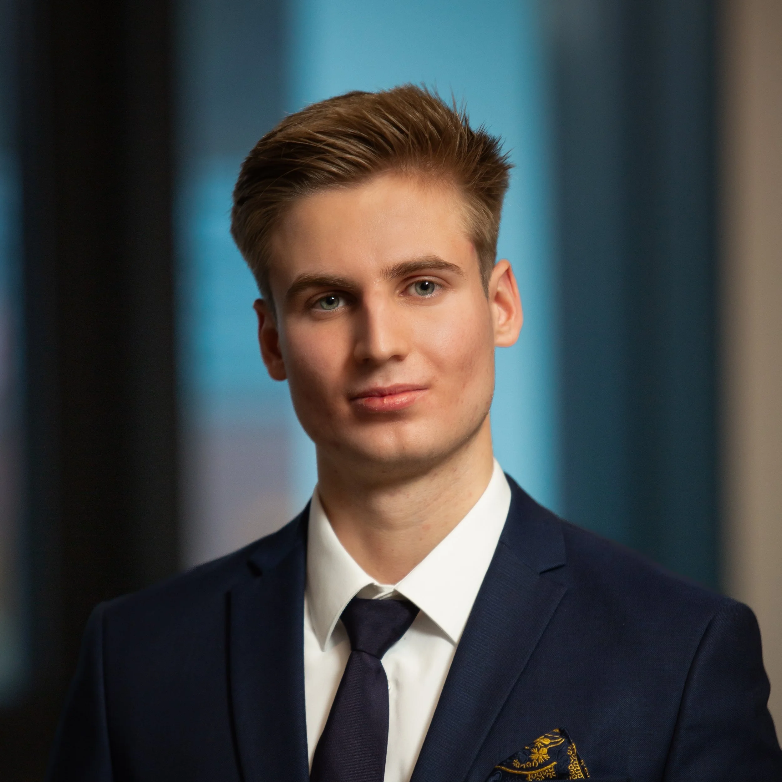 A young man in a navy blue suit, white shirt, and black tie, with a patterned pocket square, standing in front of blurred blue and black background.